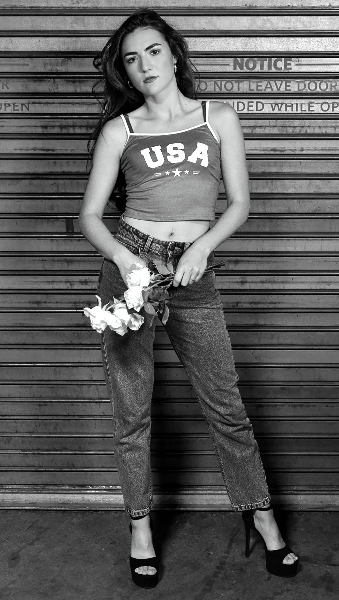 A woman with long dark hair, wearing a tank top with 'USA' on it, high heels, and jeans, standing in front of a metal door with signage, holding a bouquet of white flowers.