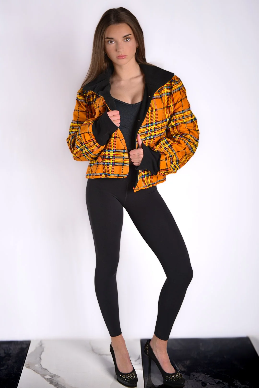 A young woman with long brown hair wearing a black top, black leggings, and high heels, holding open a yellow plaid jacket with black accents, standing against a white background.