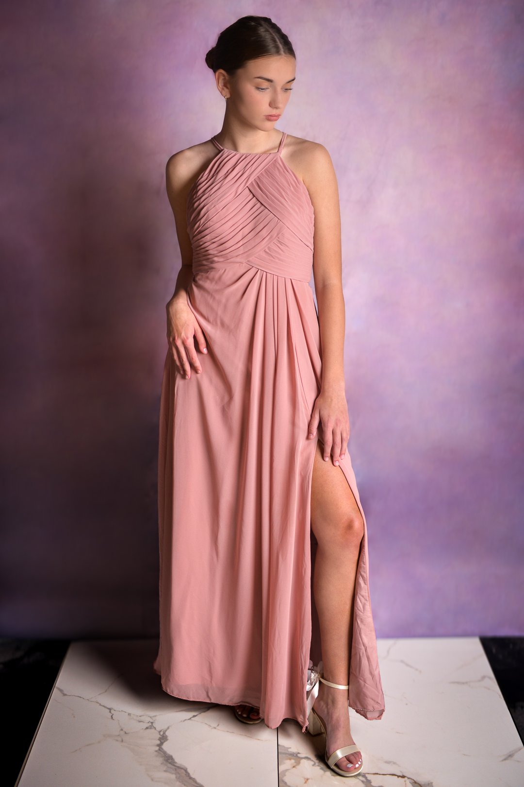 A young woman in a long, pink, pleated gown with a high slit, standing in front of a purple gradient background. She is looking down and has her hair styled in an elegant updo.