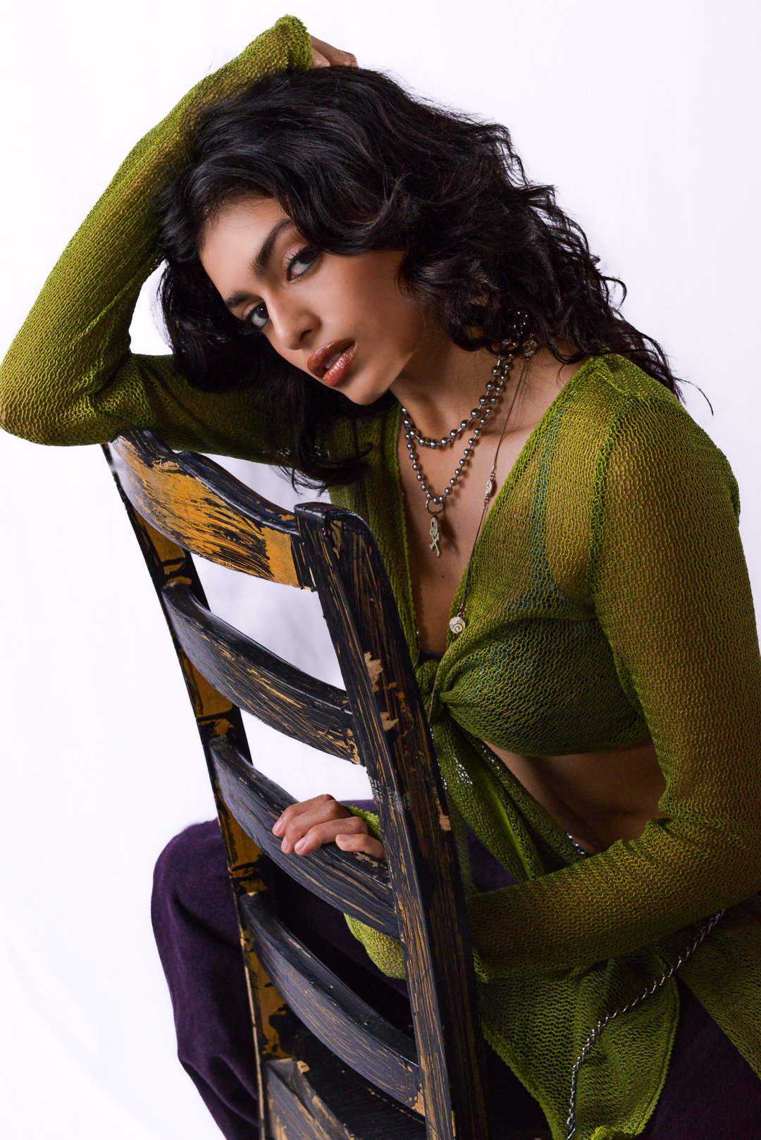 A woman with black curly hair wearing a green knit cardigan, sitting on a distressed black and yellow wooden chair, posing with one arm resting on her head, against a plain white background.