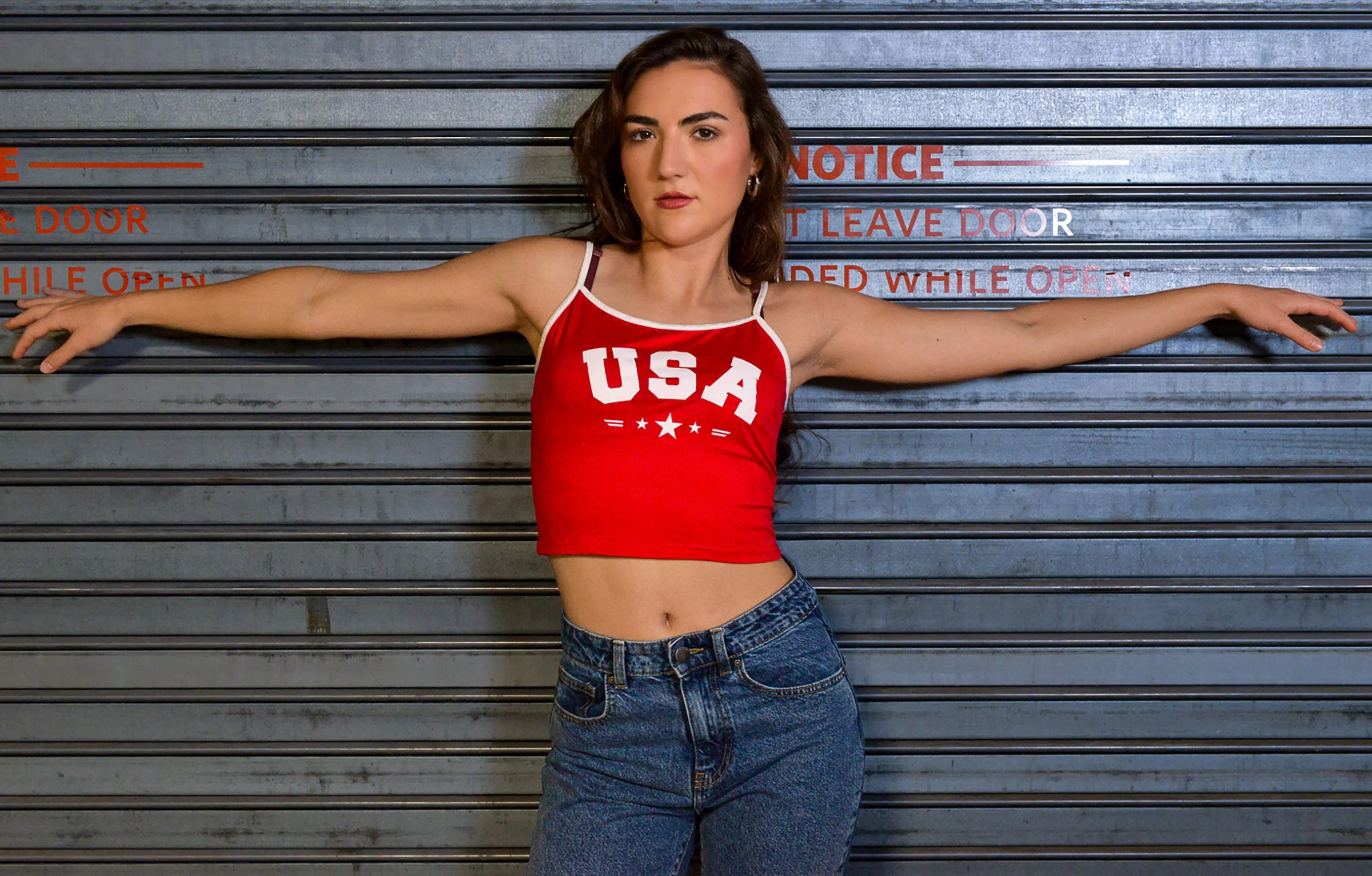 A young woman with dark hair and light skin is standing in front of a metallic garage door with red text, wearing a red tank top with the letters 'USA' and blue jeans, extending her arms out to the sides.
