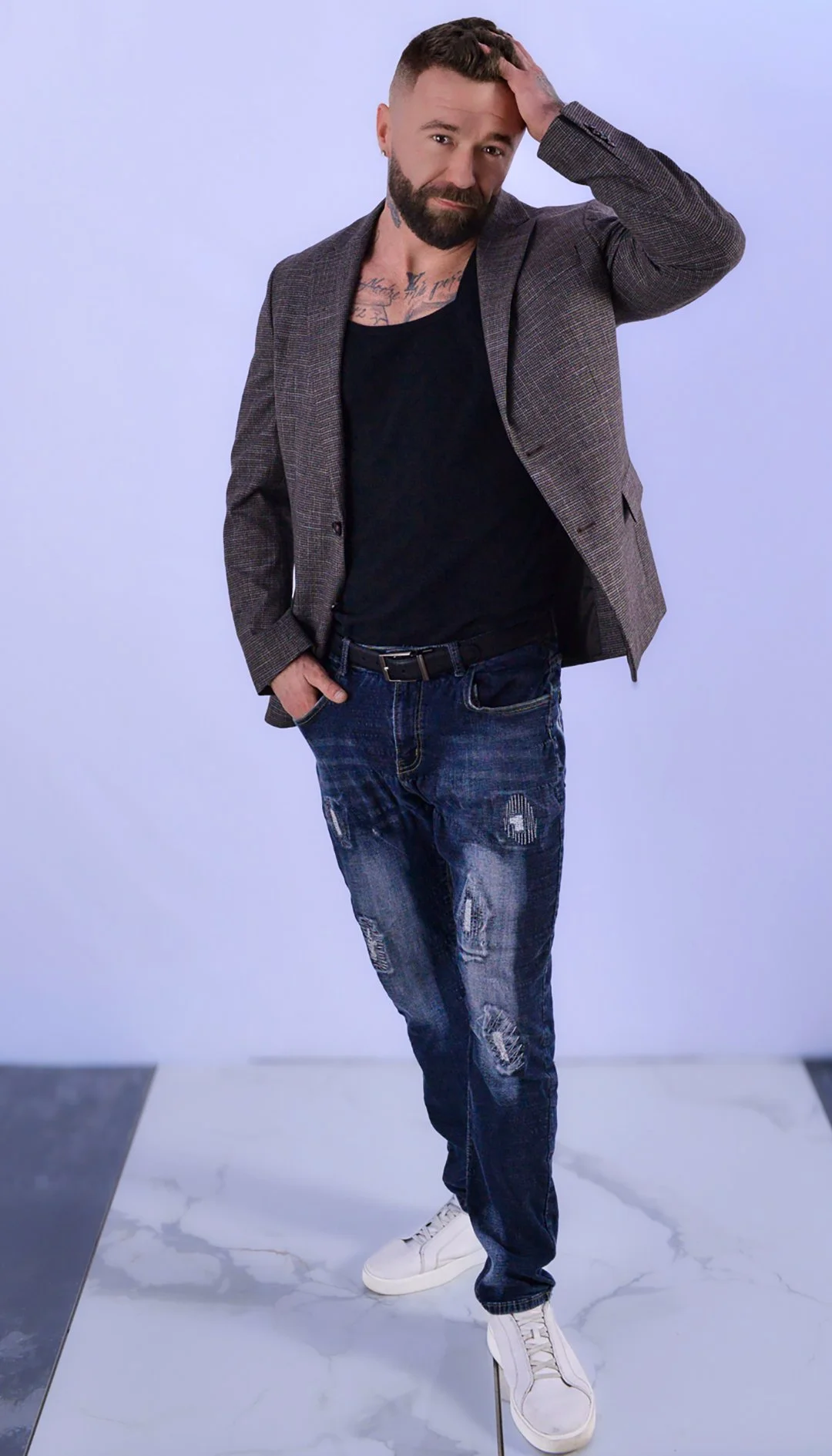 A man with a beard and tattoos on his chest, wearing a gray blazer, black t-shirt, distressed jeans, and white sneakers, standing against a plain background, with one hand on his head and the other in his pocket.