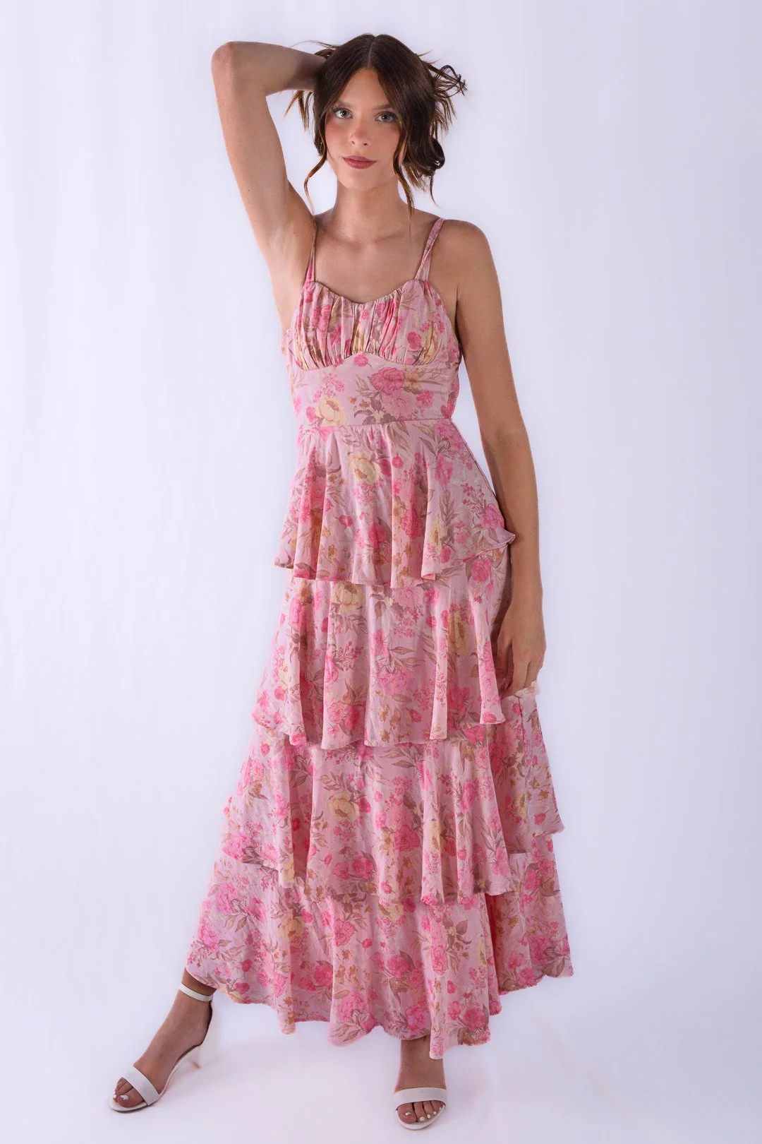 A young woman with shoulder-length brown hair wearing a pink floral tiered maxi dress and white high-heeled sandals standing against a plain white background.