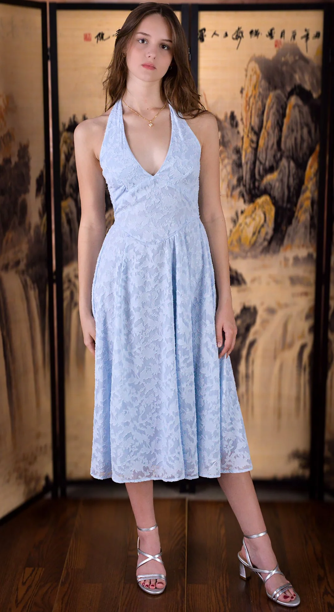 A young woman with long brown hair wearing a light blue lace halter dress, standing in front of a traditional Asian folding screen with painted landscape and Chinese calligraphy.