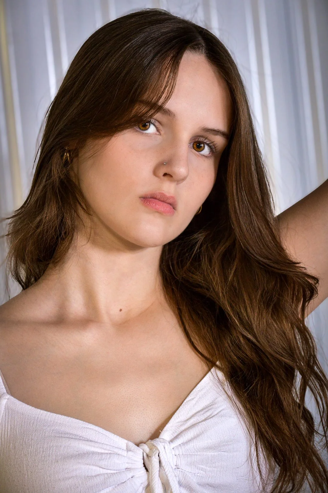 A young woman with long, wavy brown hair and brown eyes, wearing a white top with a tied knot front, stands against a lightly textured background. She has a nose piercing and small hoop earrings.