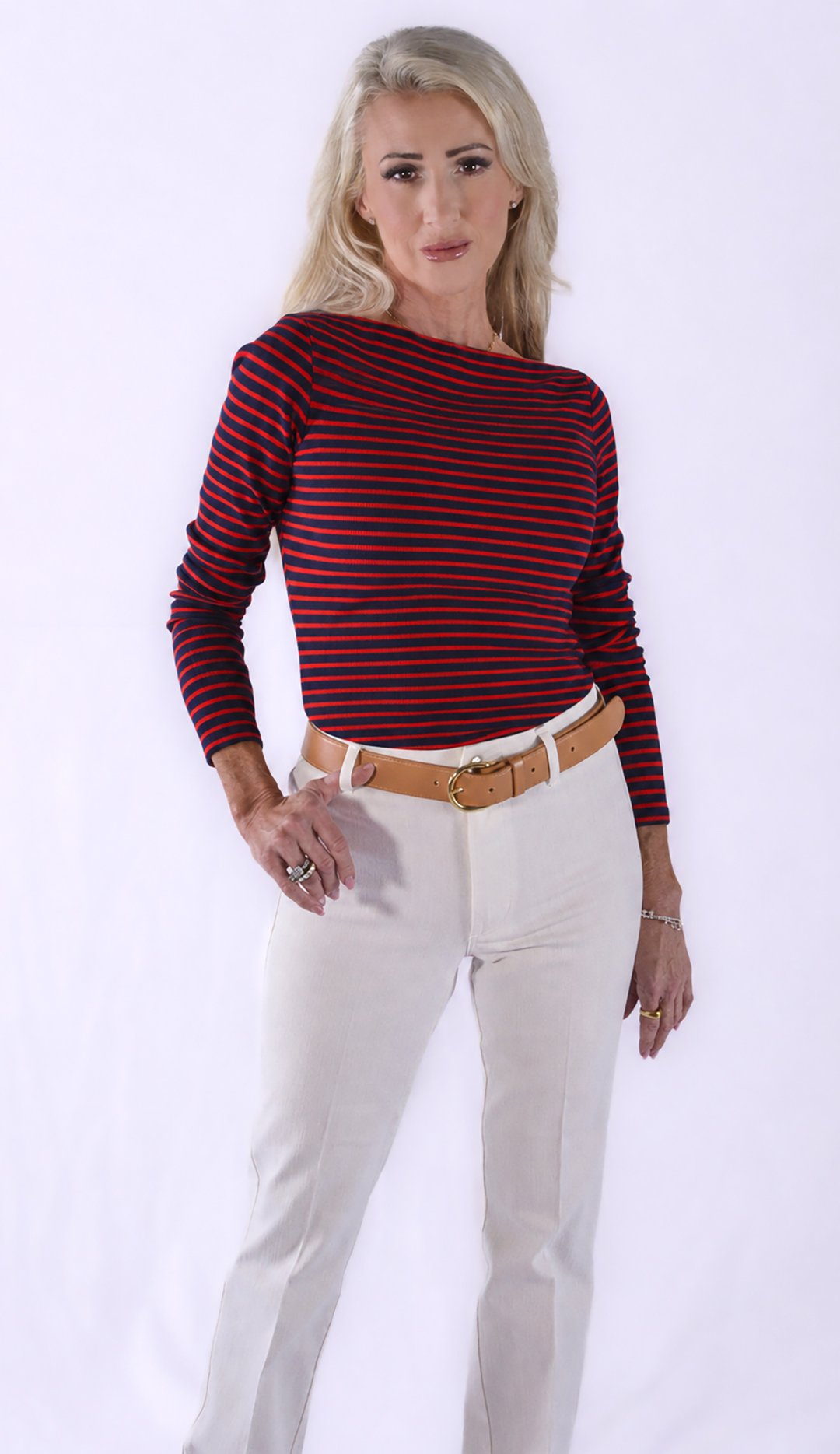 A woman with blonde hair wearing a red and navy striped long-sleeve shirt, white pants, a brown belt, and jewelry, standing against a plain white background.
