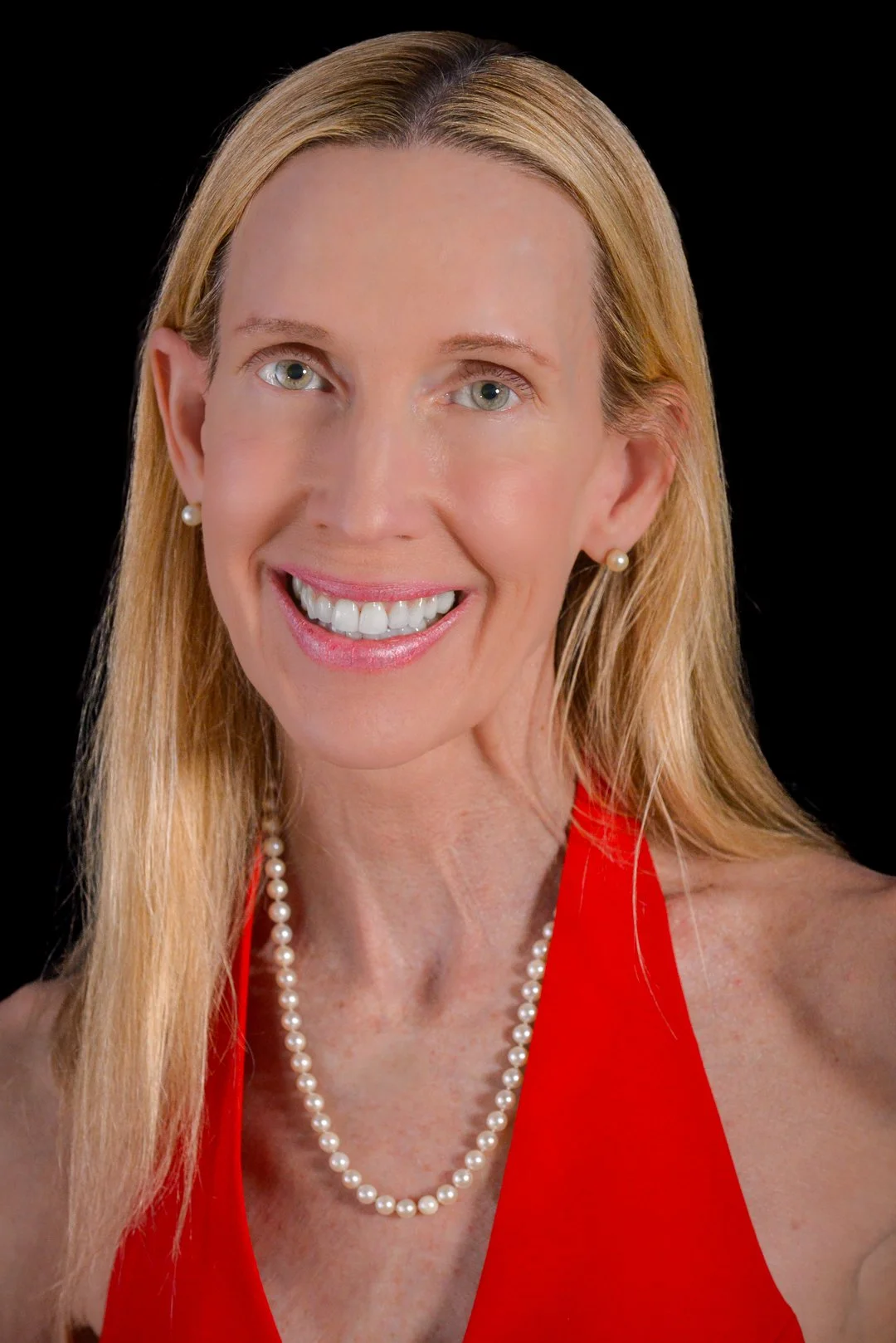 A smiling woman with long blonde hair wearing pearl earrings and necklace, a red halter top, against a black background.