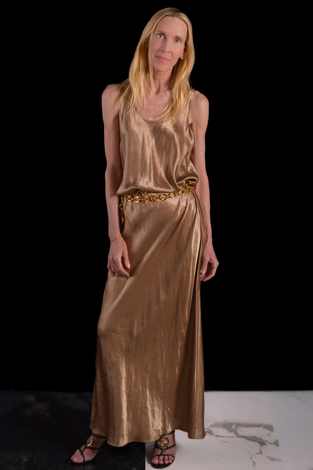 A woman with long blonde hair dressed in a shiny, gold satin sleeveless top and matching long skirt, accessorized with a chunky gold chain belt and wearing black heels with gold accents, posing against a black background.