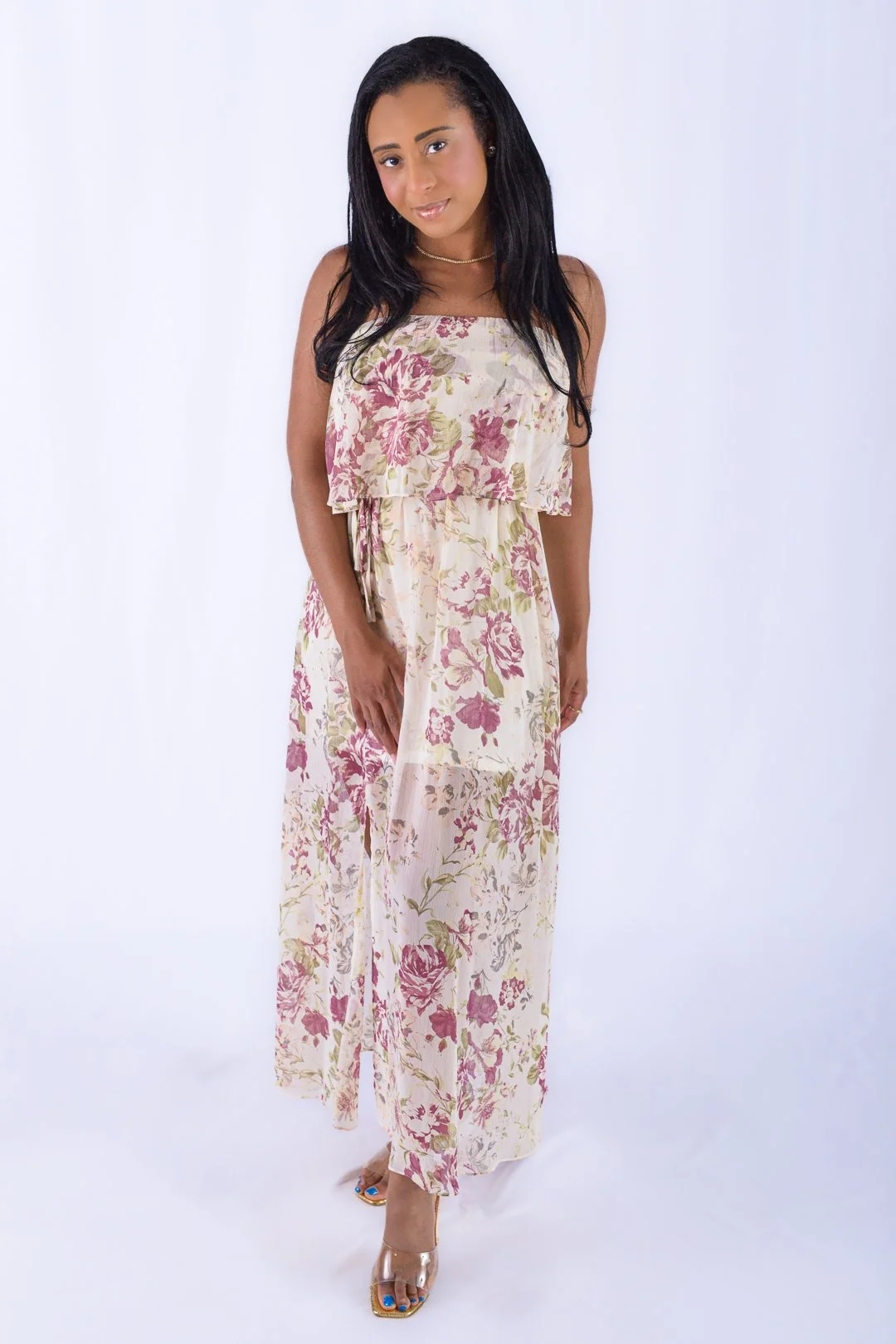 A woman in a sleeveless, floral maxi dress with pink and green flowers on a cream background, standing against a plain white backdrop.