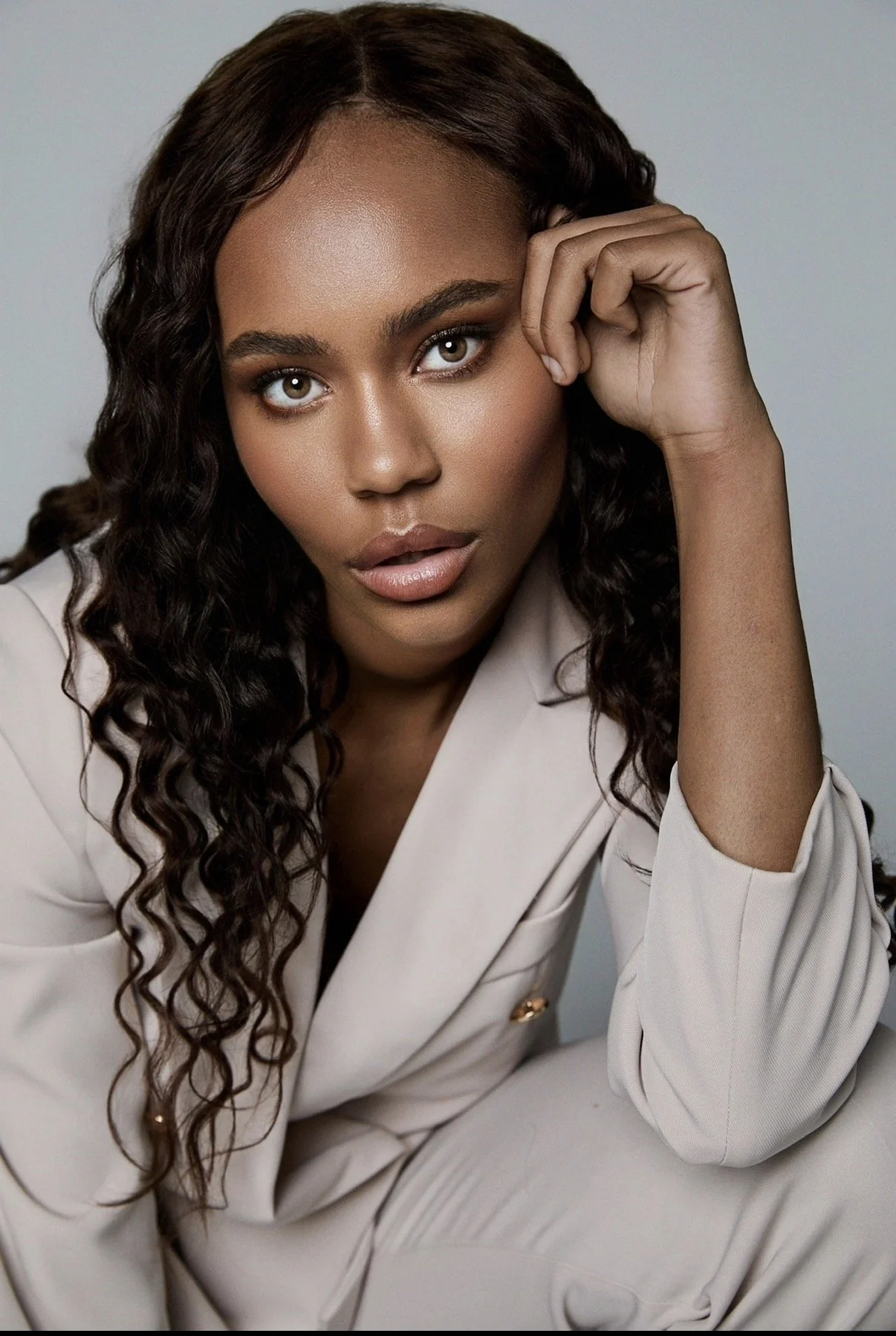 Portrait of a woman with long curly hair and brown skin, wearing a beige suit, posing with her right hand near her face against a plain gray background.