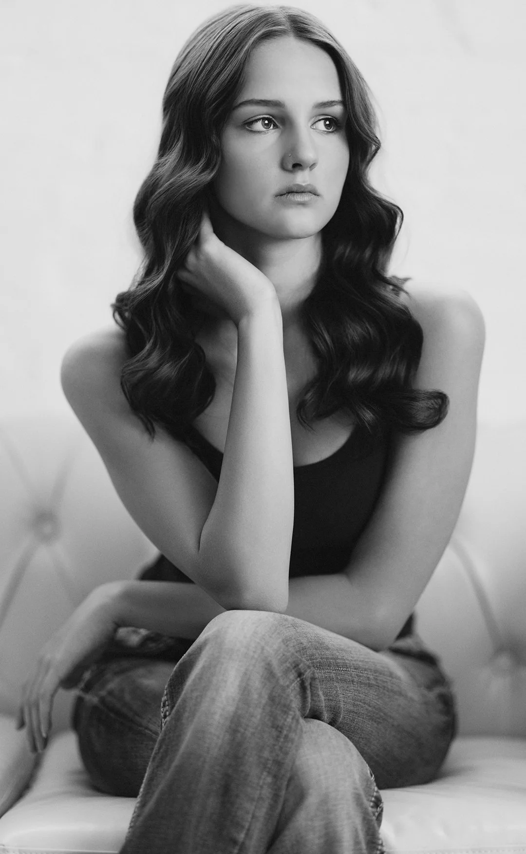 Black and white photograph of a young woman with long wavy hair, sitting on a bed, resting her chin on her hand, wearing a tank top and jeans.