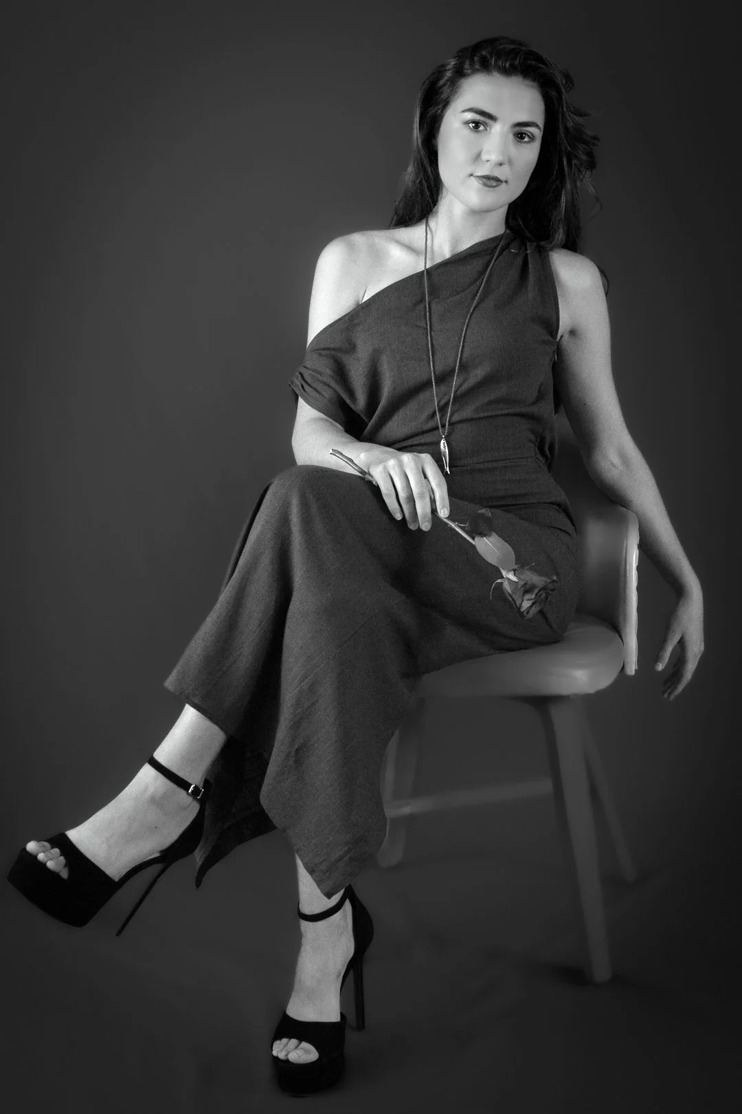 A black and white photo of a woman sitting on a chair, wearing a sleeveless dress, high heels, and jewelry, with a confident expression.