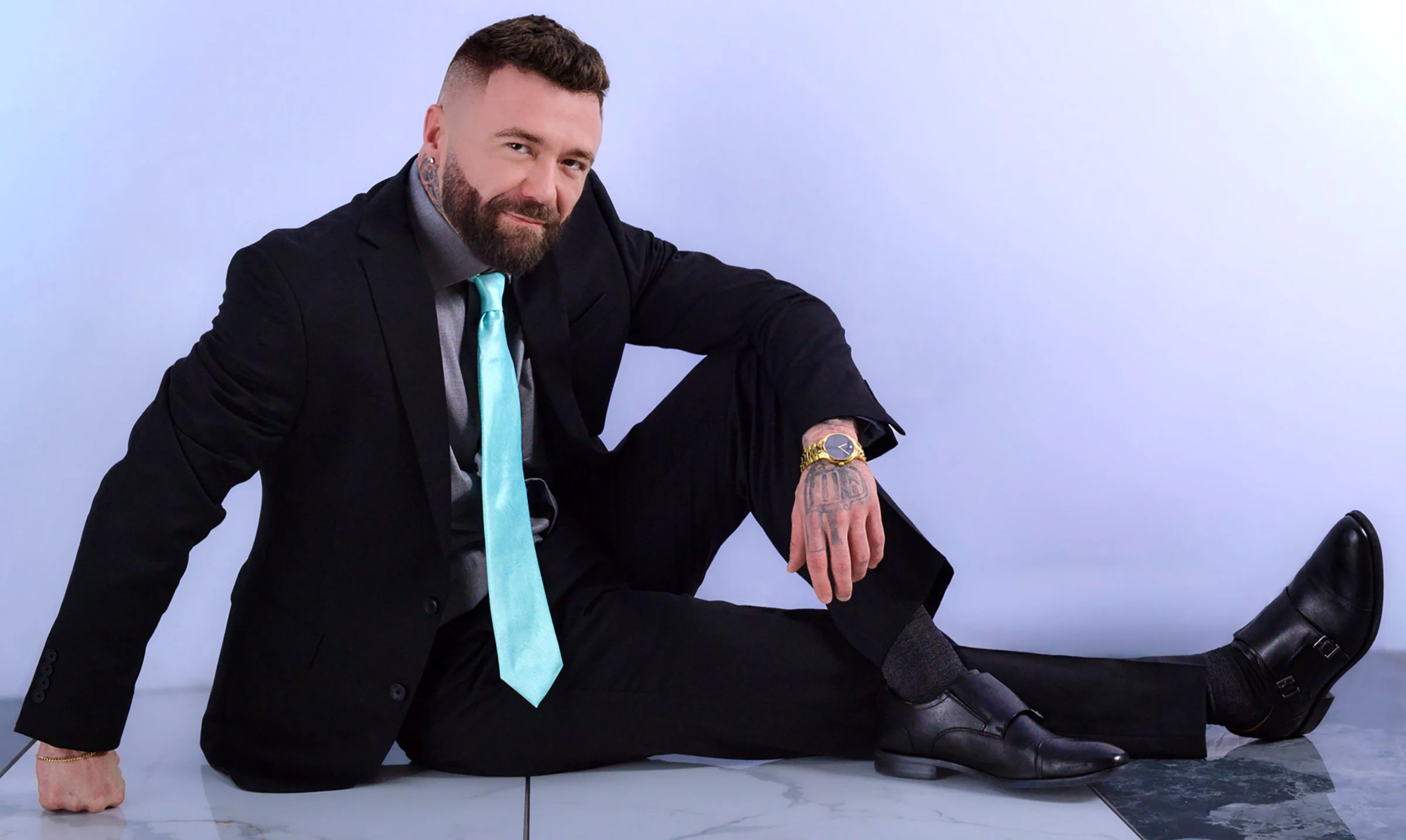A bearded man with tattoos and a gold watch, dressed in a black suit with a gray shirt and a light blue tie, is sitting on the floor with his left leg extended and his right knee bent. He is leaning on his left hand and has his right hand resting on 