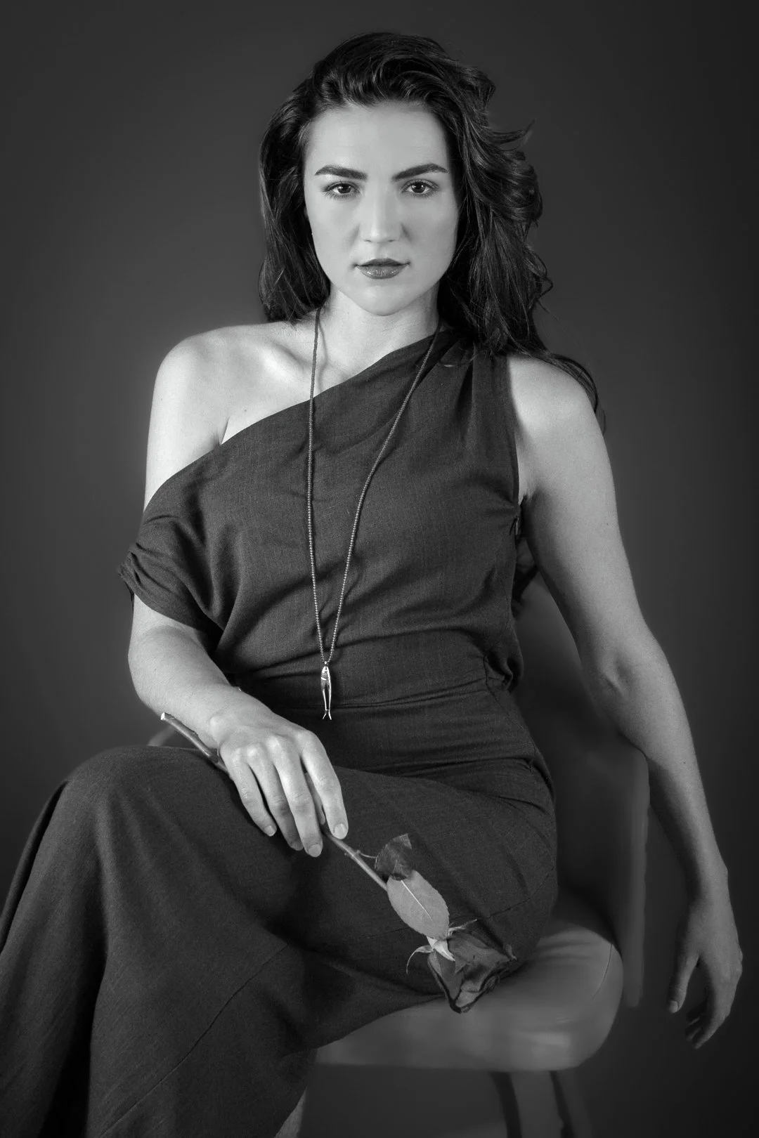A woman with dark hair and a serious expression, sitting on a chair, holding a rose in her hand, dressed in a sleeveless top and long skirt, in a black-and-white studio portrait.