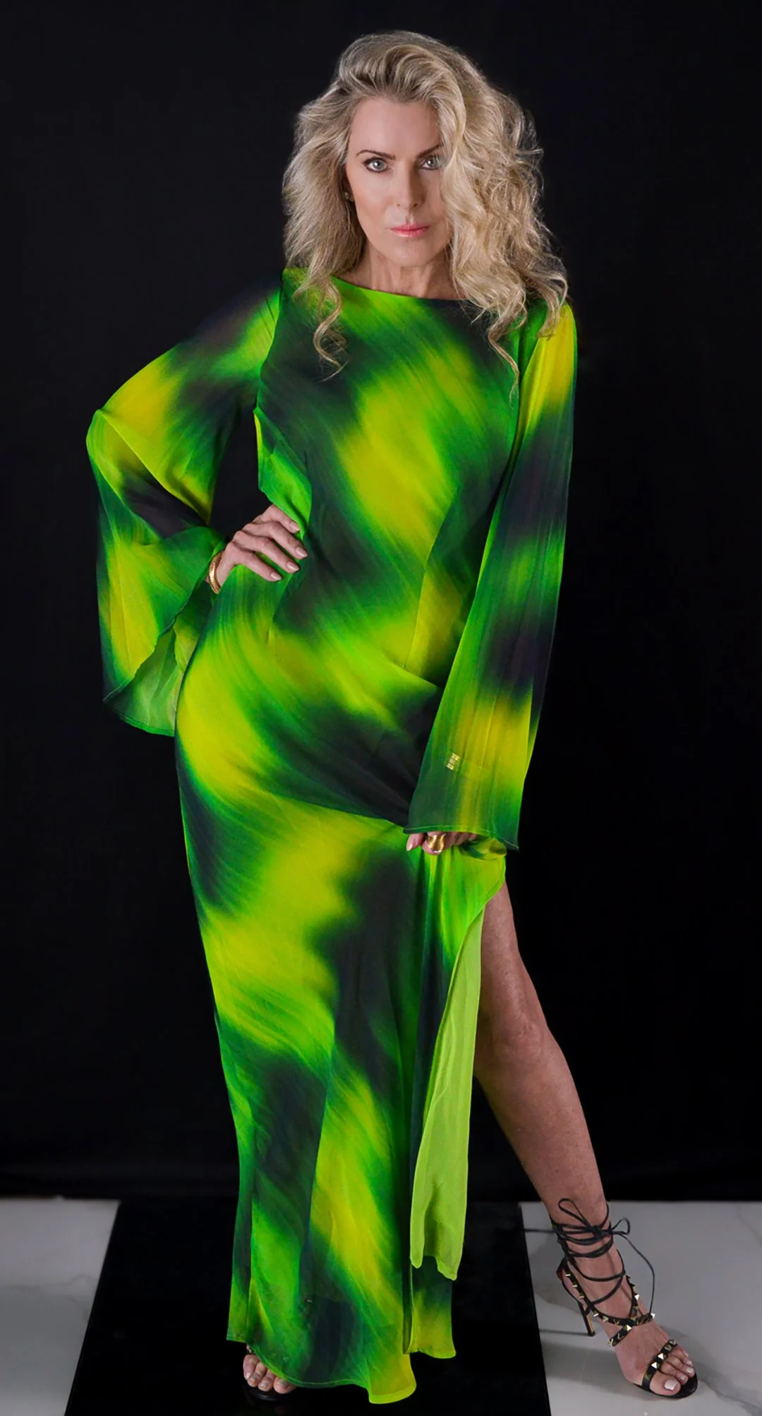 Woman with blonde, wavy hair wearing a flowing, neon green and black patterned dress with a thigh-high slit, standing on a black runway against a black background.