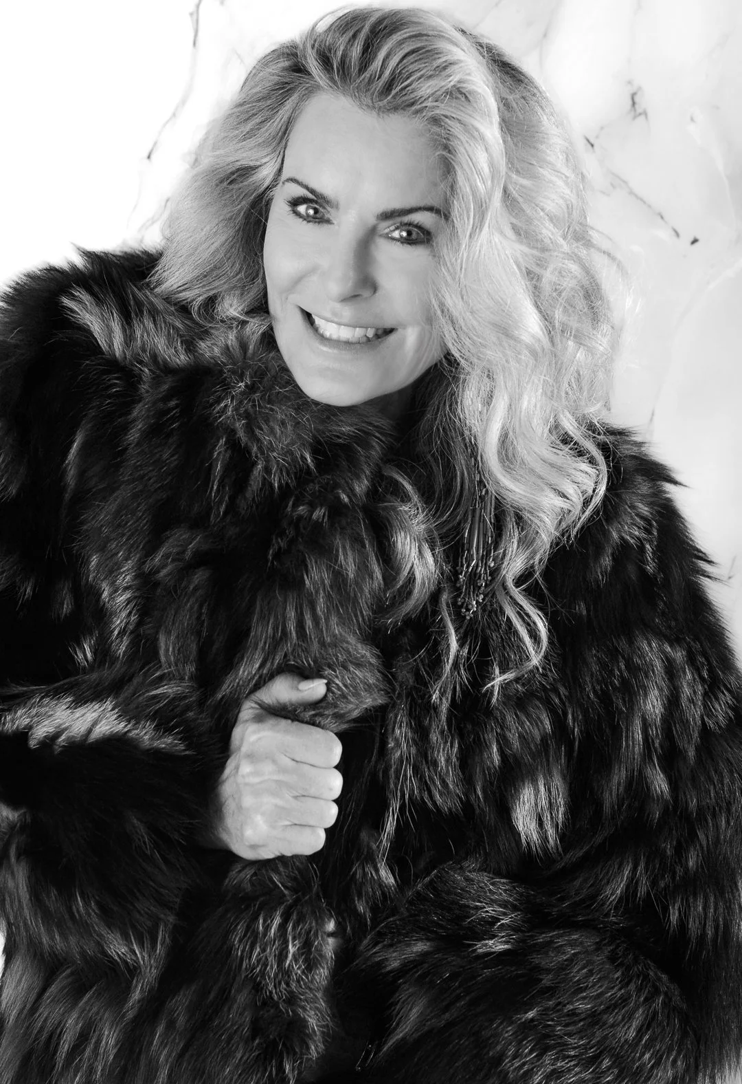 A woman with wavy blonde hair smiling and wearing a dark fur coat, holding a fur garment or accessory, in black and white.
