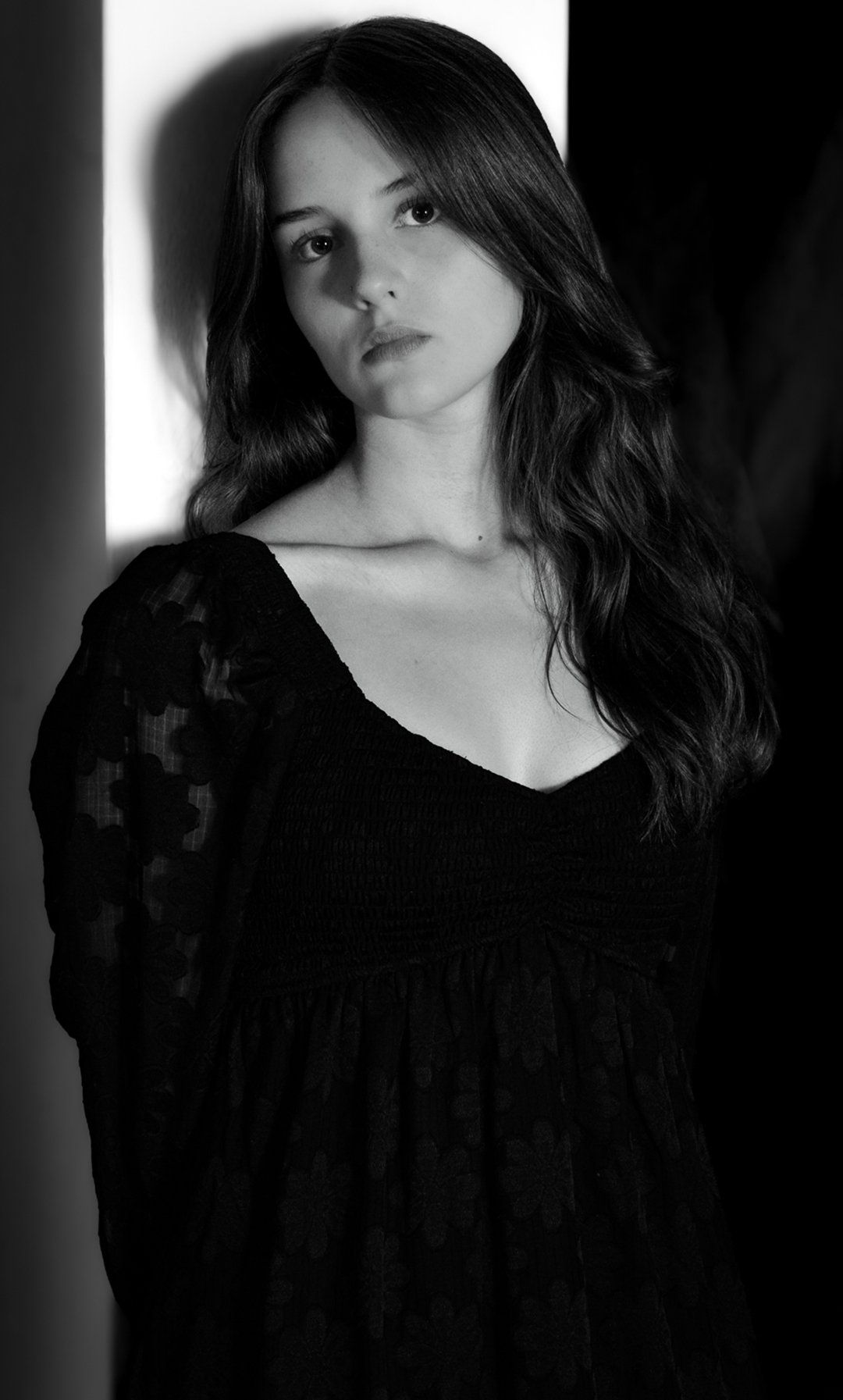 Black and white portrait of a young woman with long wavy hair, wearing a dark dress with a textured bodice and sheer sleeves with floral accents, standing against a light and dark background.
