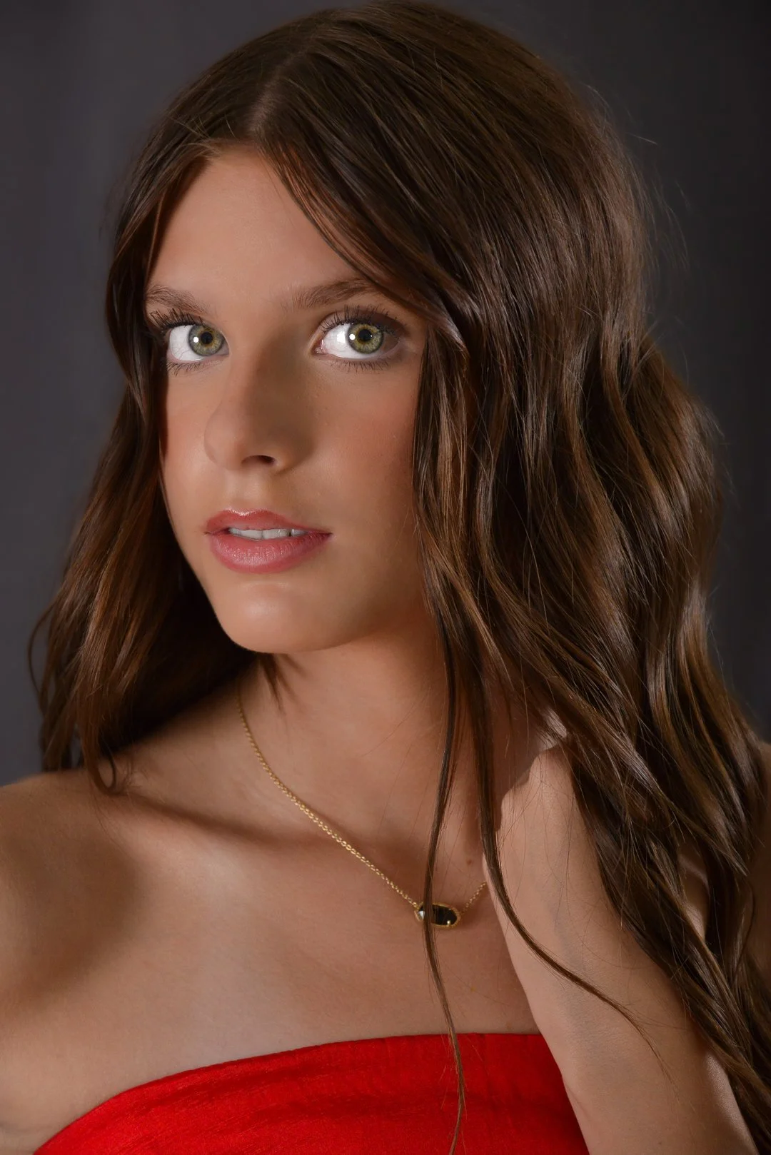 A young woman with long, wavy brown hair, green eyes, and light makeup wearing a red strapless top and a delicate gold necklace, against a dark background.