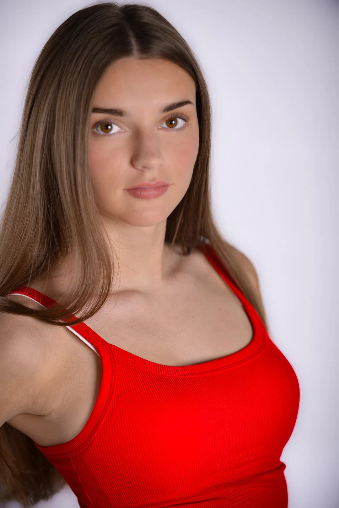 A young woman wearing a red tank top with long brown hair, standing against a plain light background.