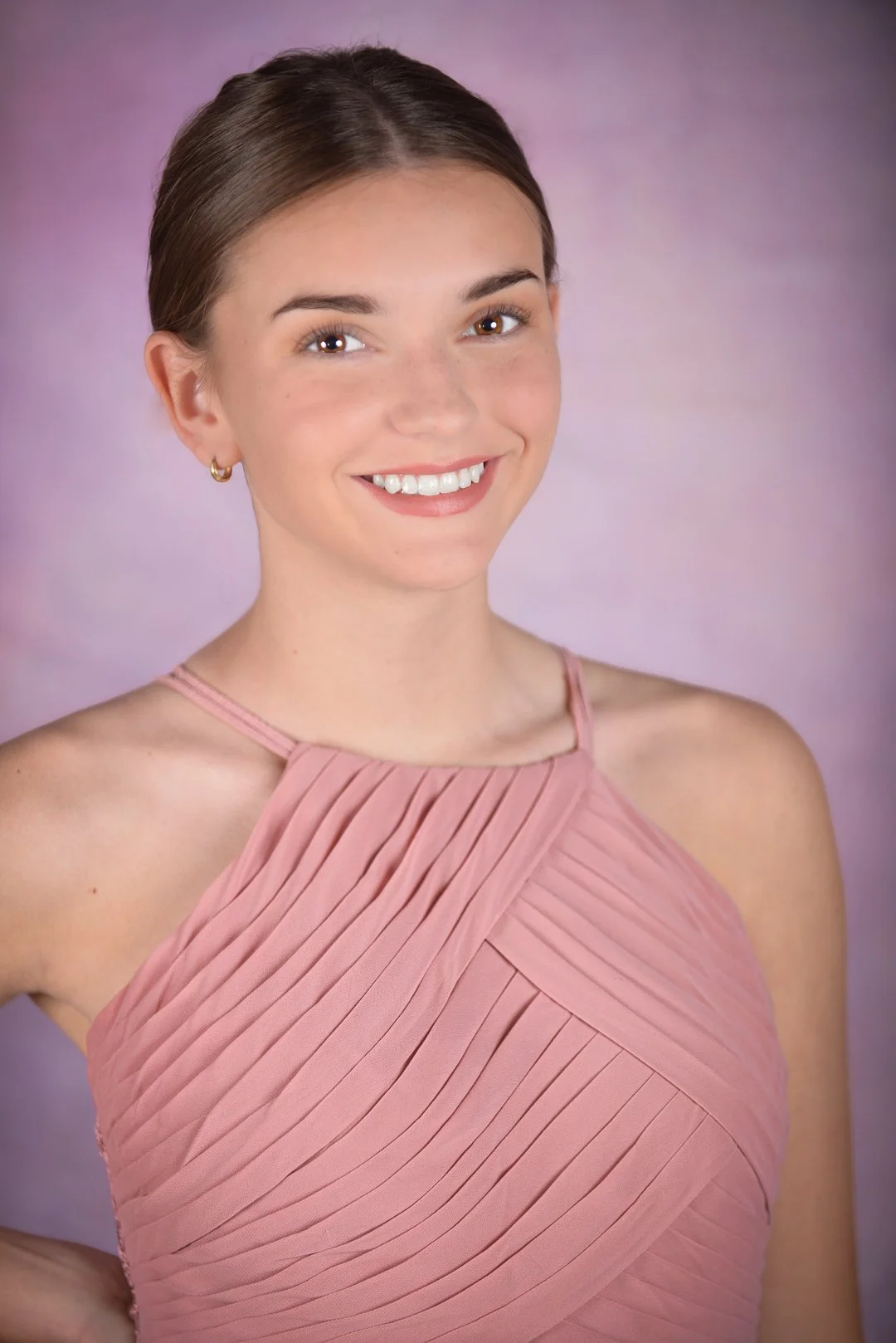 A young woman with brown hair styled in a bun, smiling, wearing a pink pleated dress, in front of a purple background.