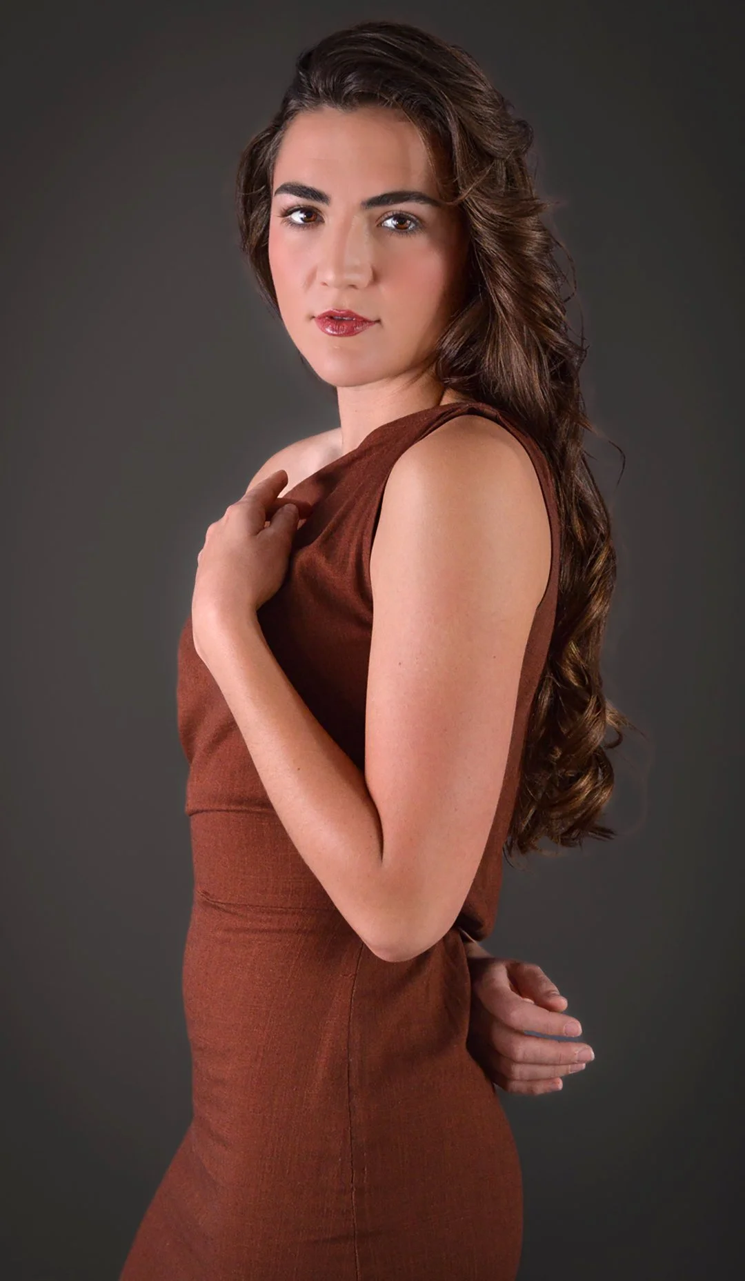A woman with long brown curly hair wearing a sleeveless brown dress, standing against a gray background, looking at the camera with a neutral expression.