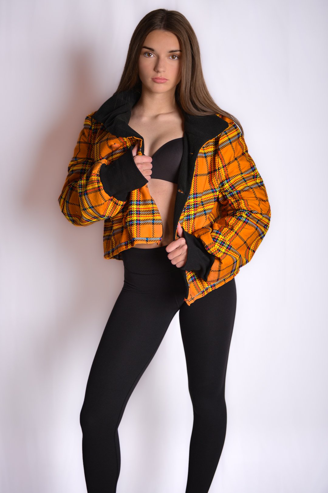 A young woman with long brown hair wearing a black bra and high-waisted black leggings, partially unzipping an orange plaid jacket with black accents, standing against a plain white wall.