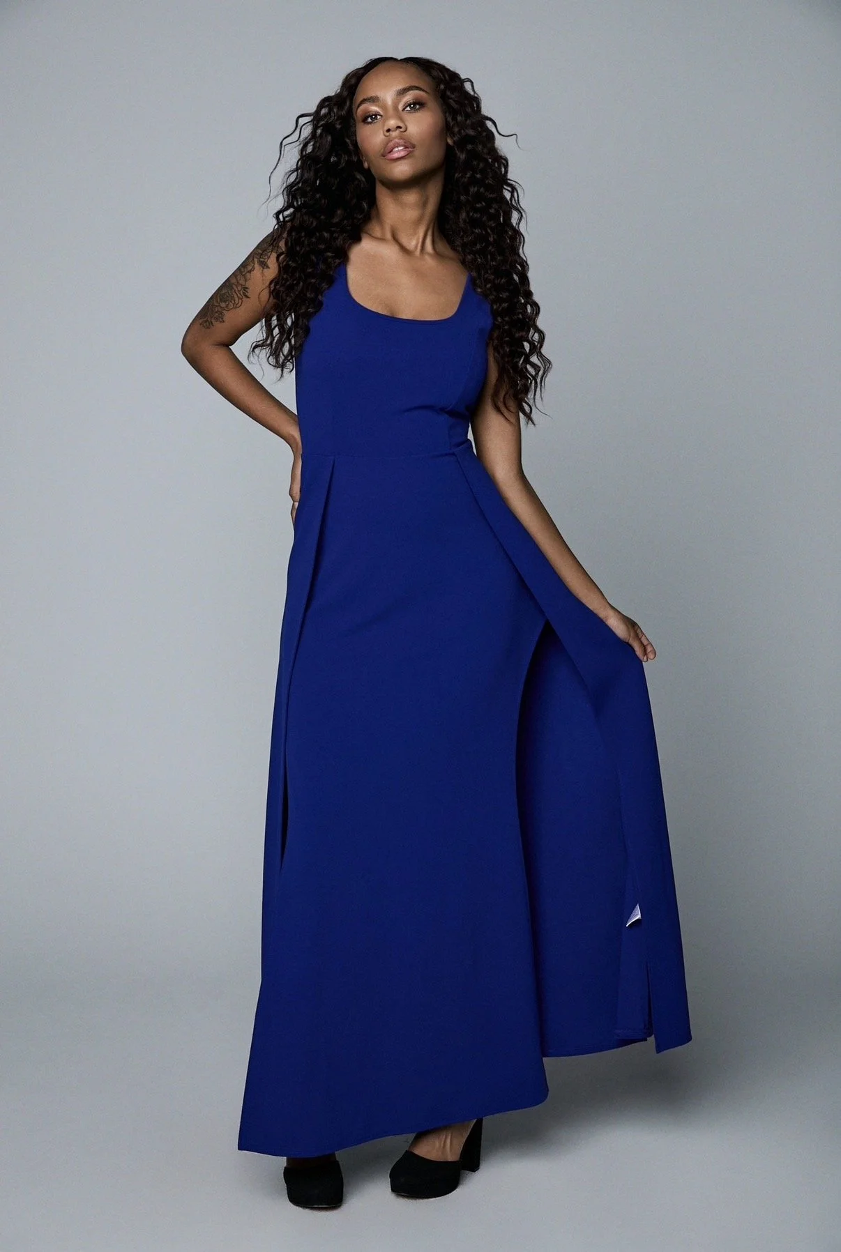 A woman with long curly black hair, wearing a sleeveless royal blue dress that reaches the floor, standing against a plain gray background. She has one hand on her hip and the other near her thigh, wearing black heeled shoes.