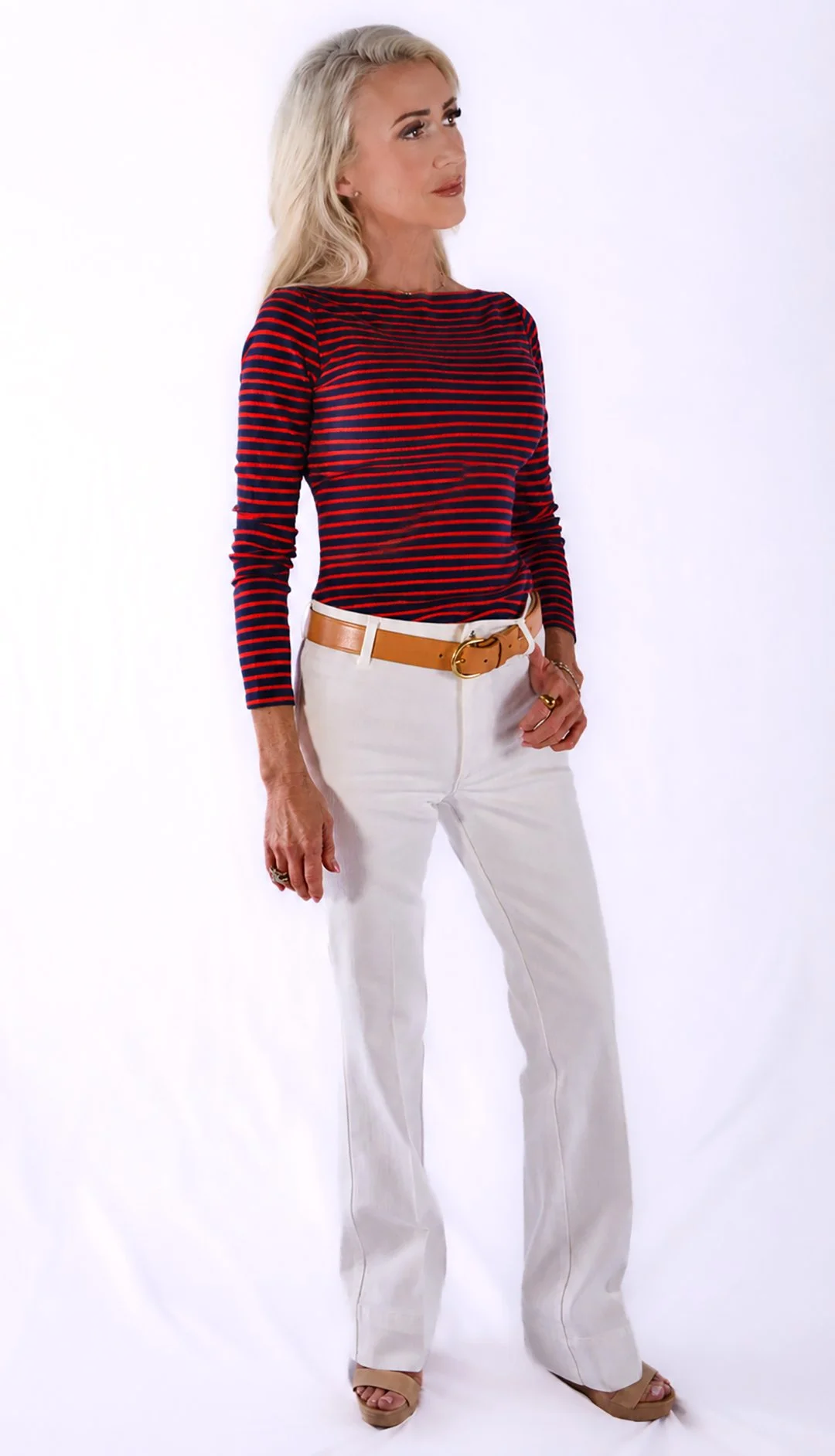 A woman with long blonde hair, wearing a red and navy striped long-sleeve shirt, white pants, a tan belt, and beige high heels, stands against a white background.