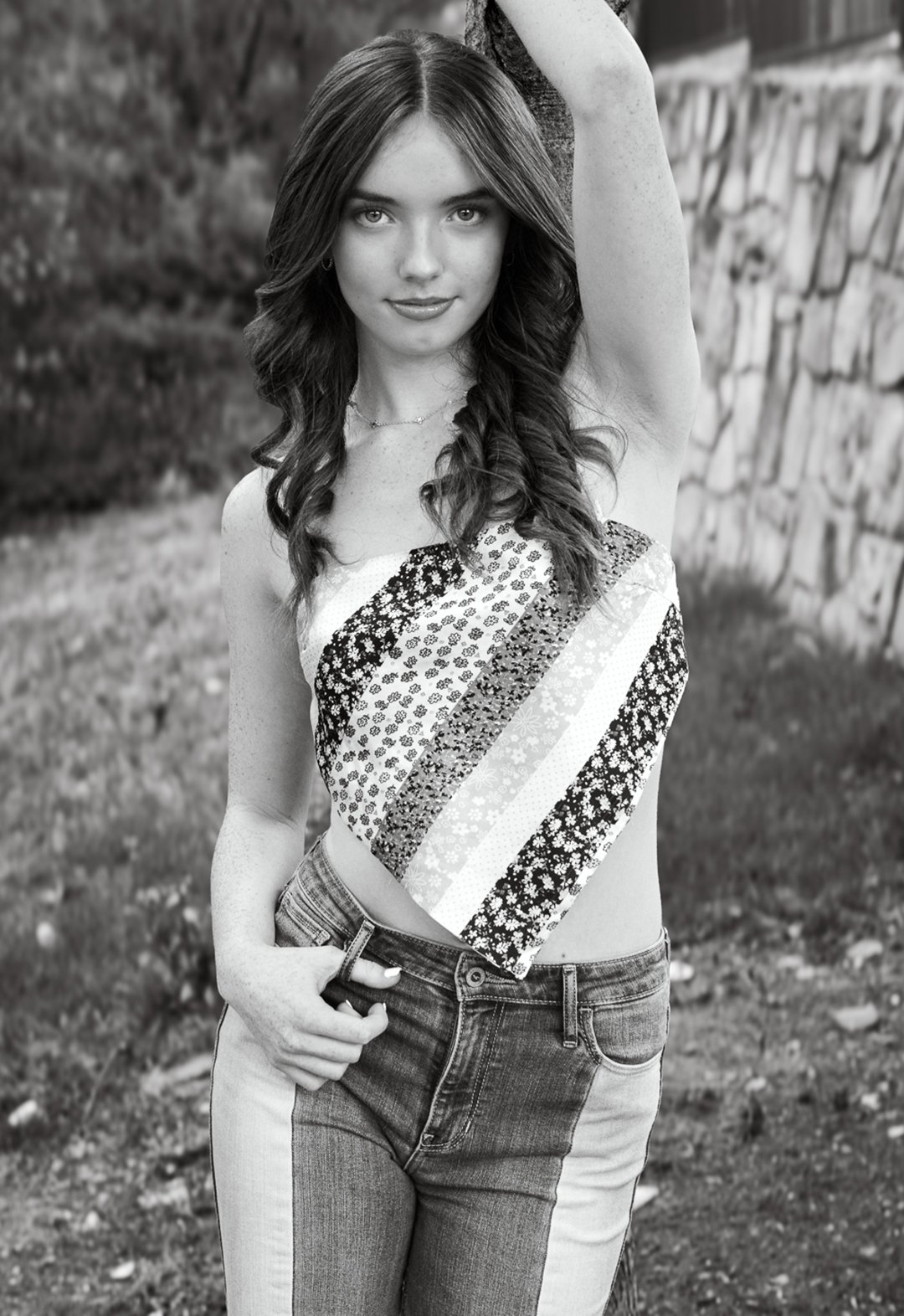 A young woman with shoulder-length wavy hair and light eyes standing outdoors with a stone wall and trees in the background. She is wearing a strapless patchwork top with floral patterns and high-waisted jeans, with one hand in her pocket and the oth