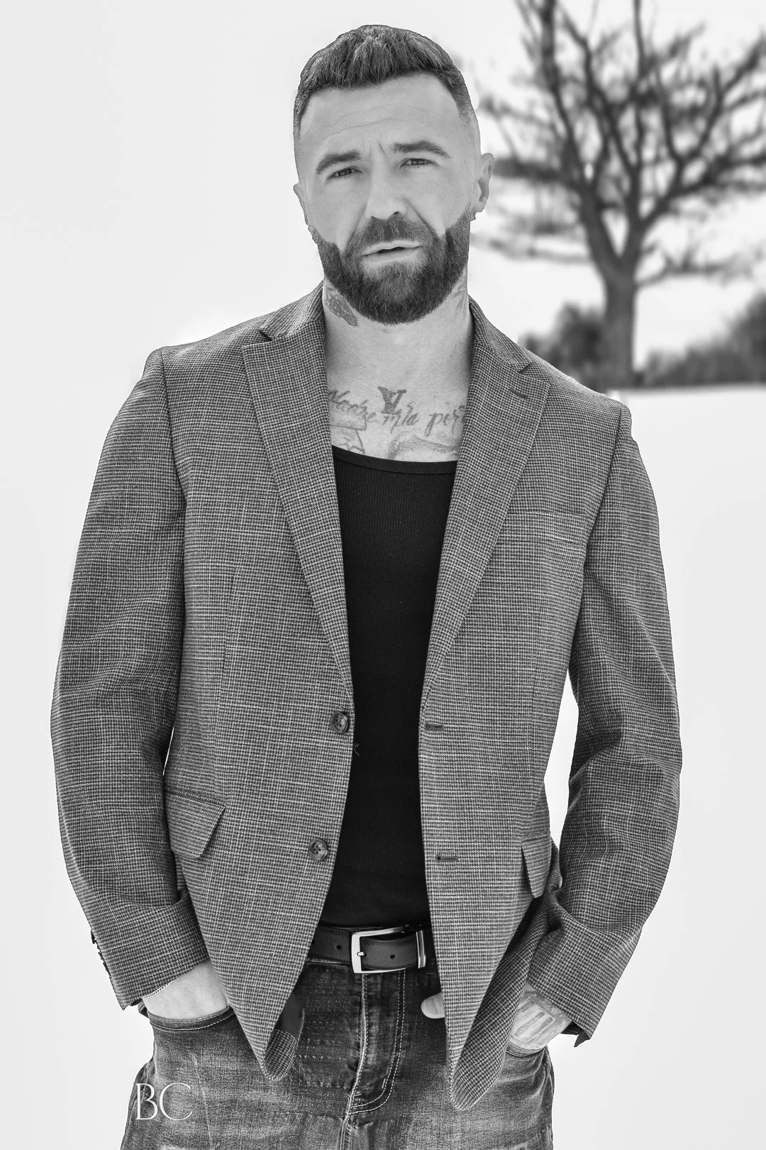 Black and white photo of a man with a beard, wearing a checkered blazer over a black t-shirt, standing outdoors with a large leafless tree in the background.