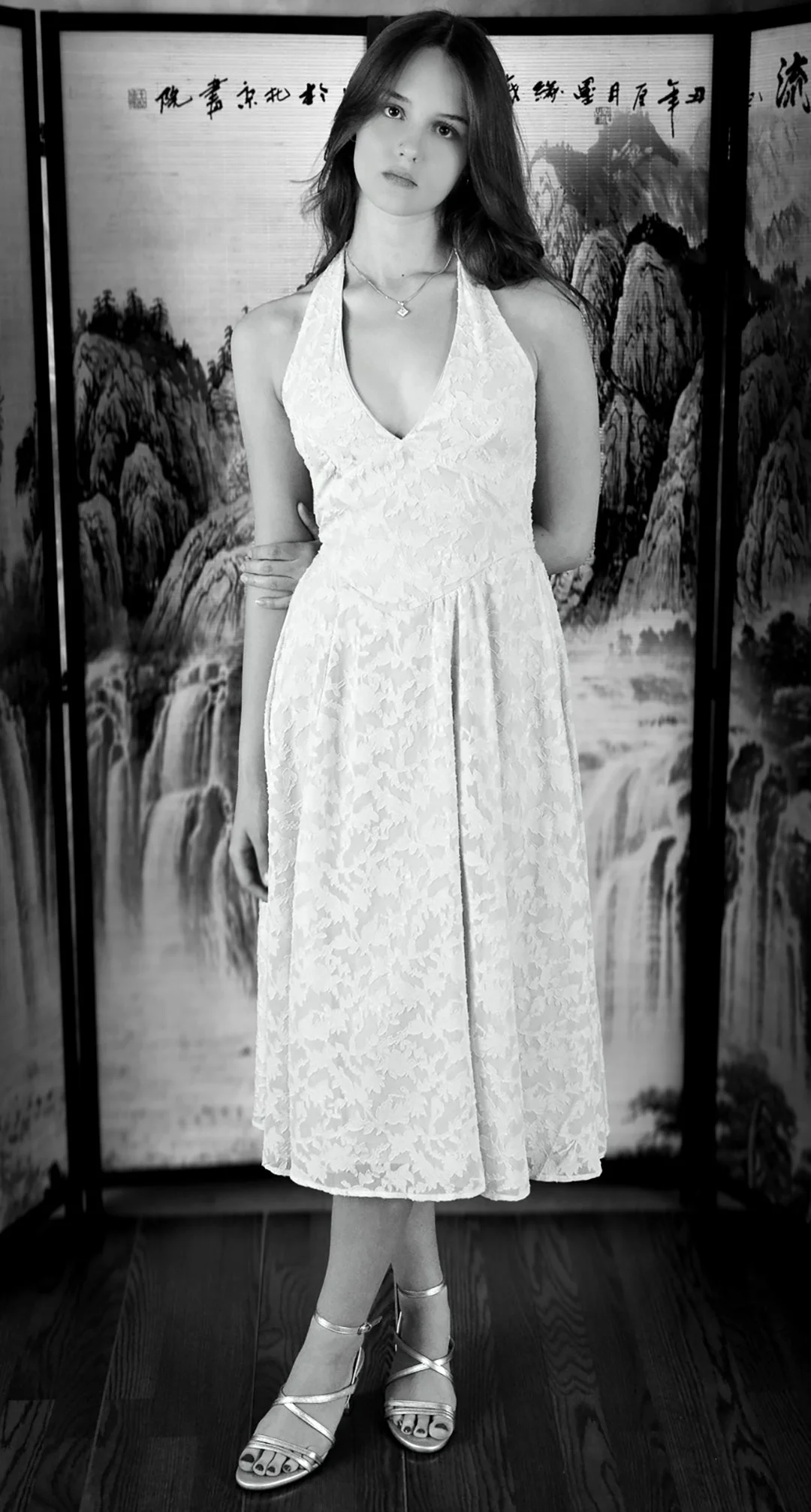 A woman in a white lace dress wearing strappy heels standing in front of a traditional Chinese landscape painting backdrop.