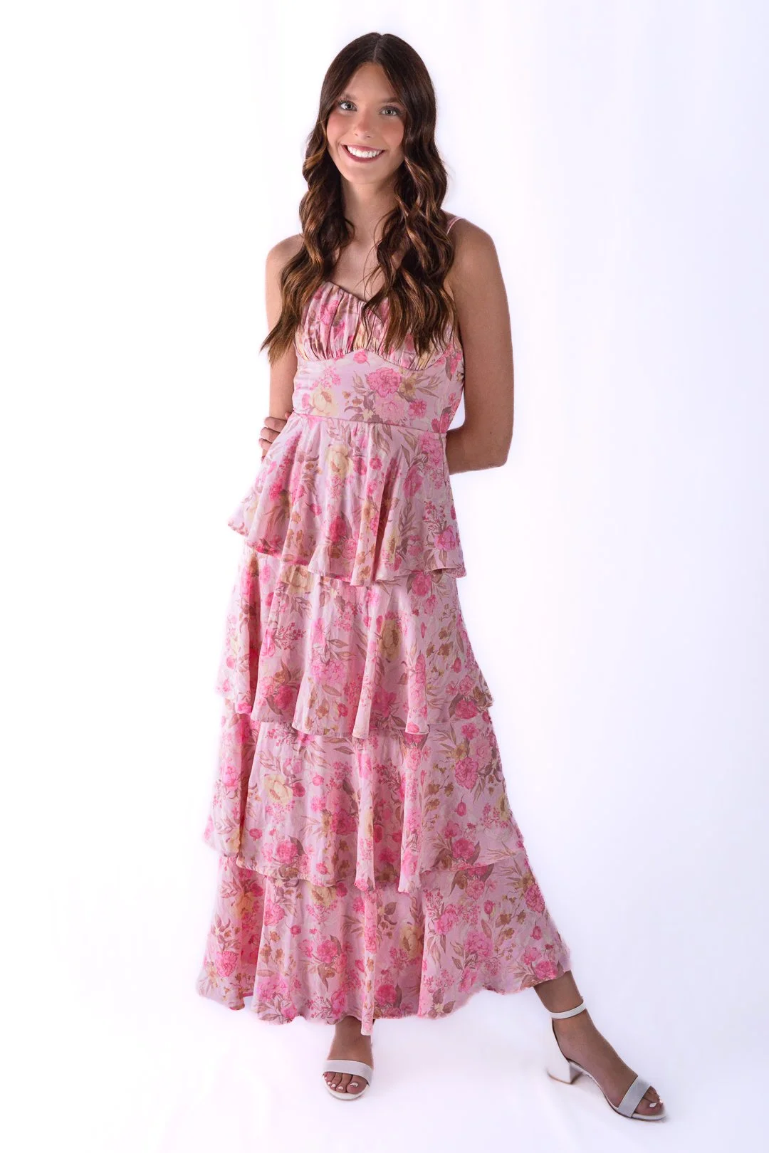 A woman with long wavy brown hair wearing a pink floral tiered maxi dress and white high-heeled sandals, standing against a white background, smiling at the camera.