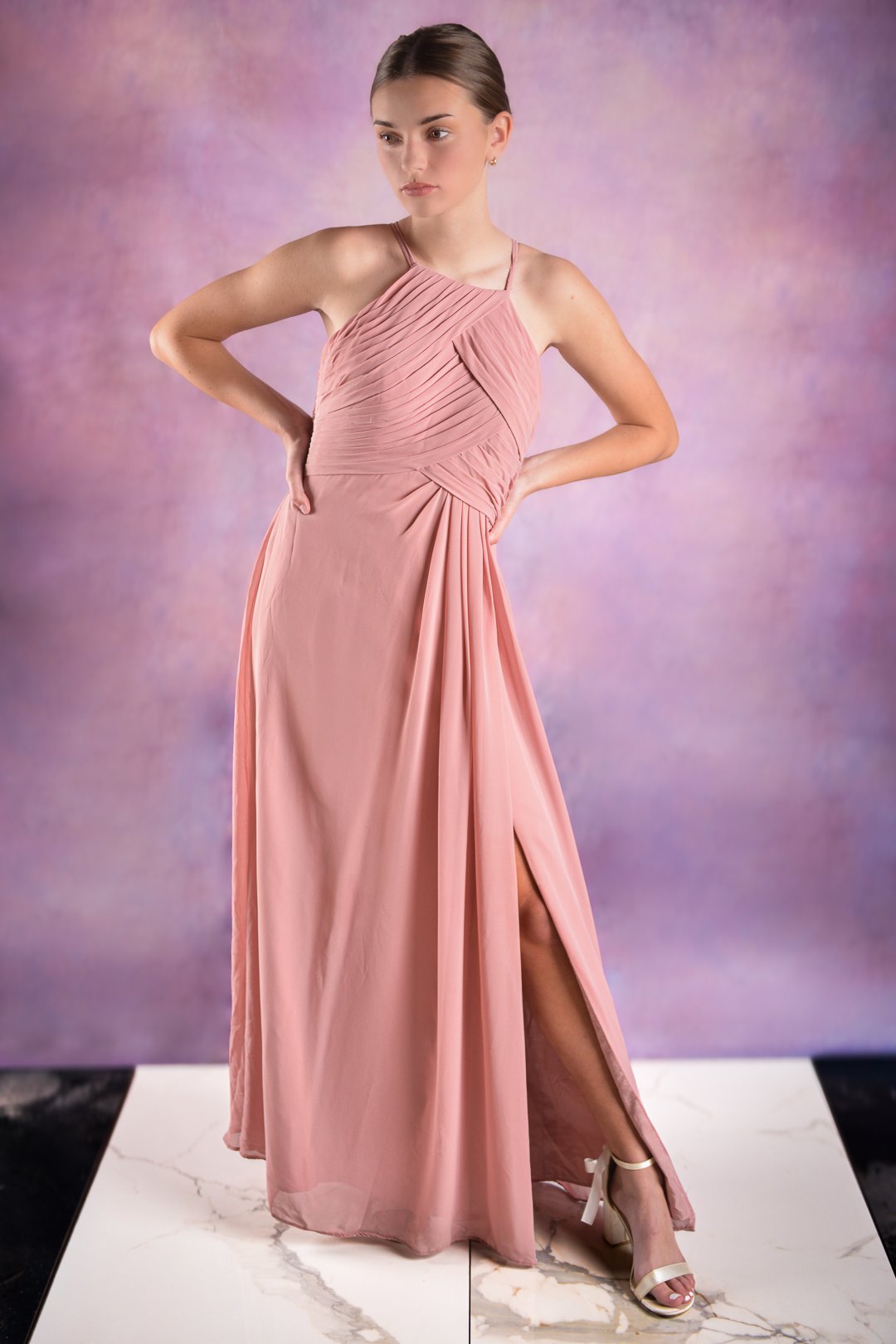 A woman in a pink pleated floor-length dress with a slit, standing on a marble platform against a purple background.