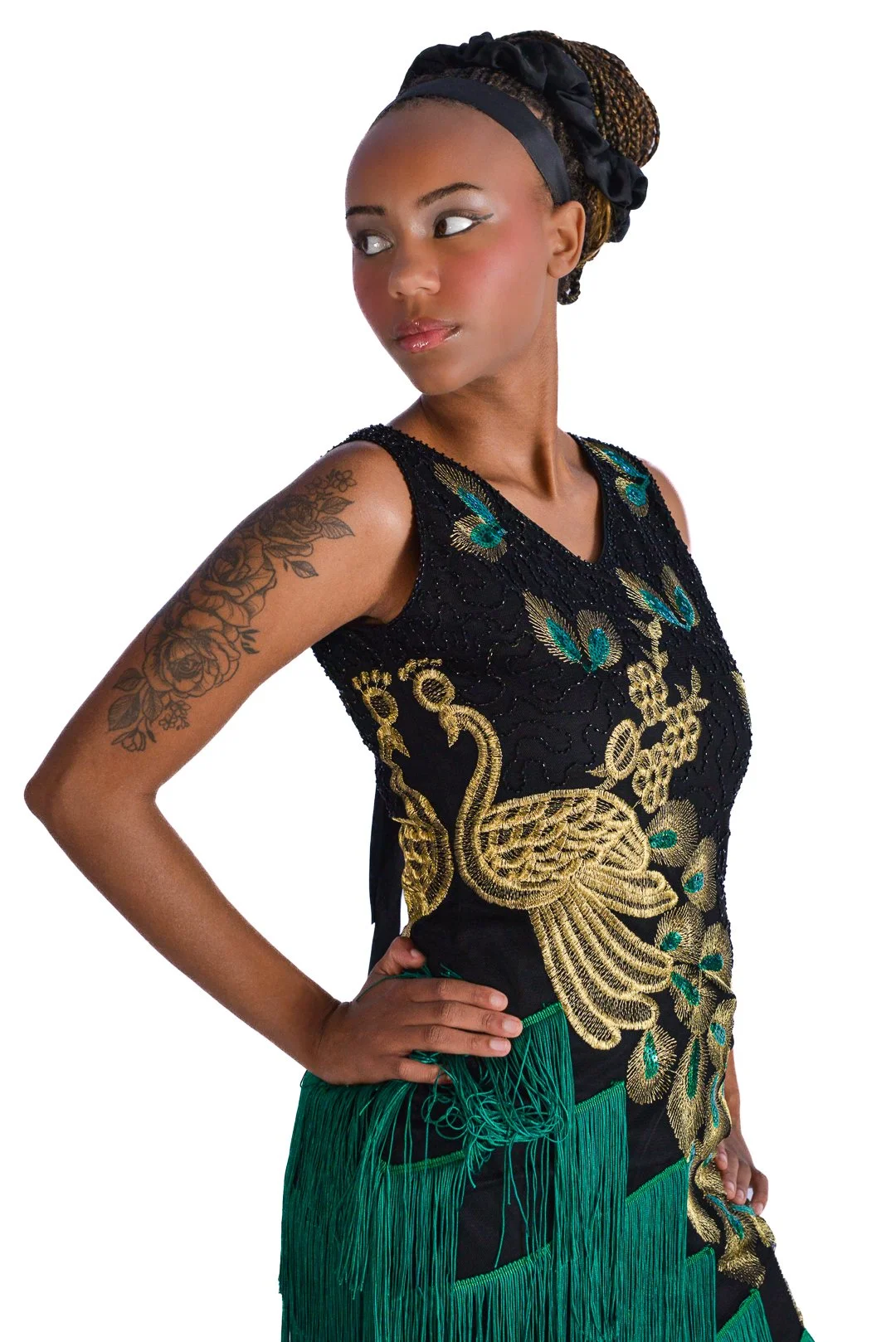 Woman with braided hair in a black head wrap, wearing a sleeveless black dress with gold and green embroidery featuring a swan and floral design, and green fringed fabric at the waist, standing with one hand on her hip.