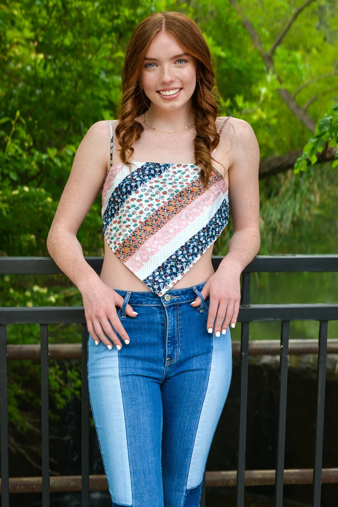 A young woman with red hair, blue eyes, and freckles, smiling outdoors. She is wearing a sleeveless, patchwork crop top with diagonal stripes in various floral patterns and high-waisted blue jeans, standing in front of a green, wooded background.