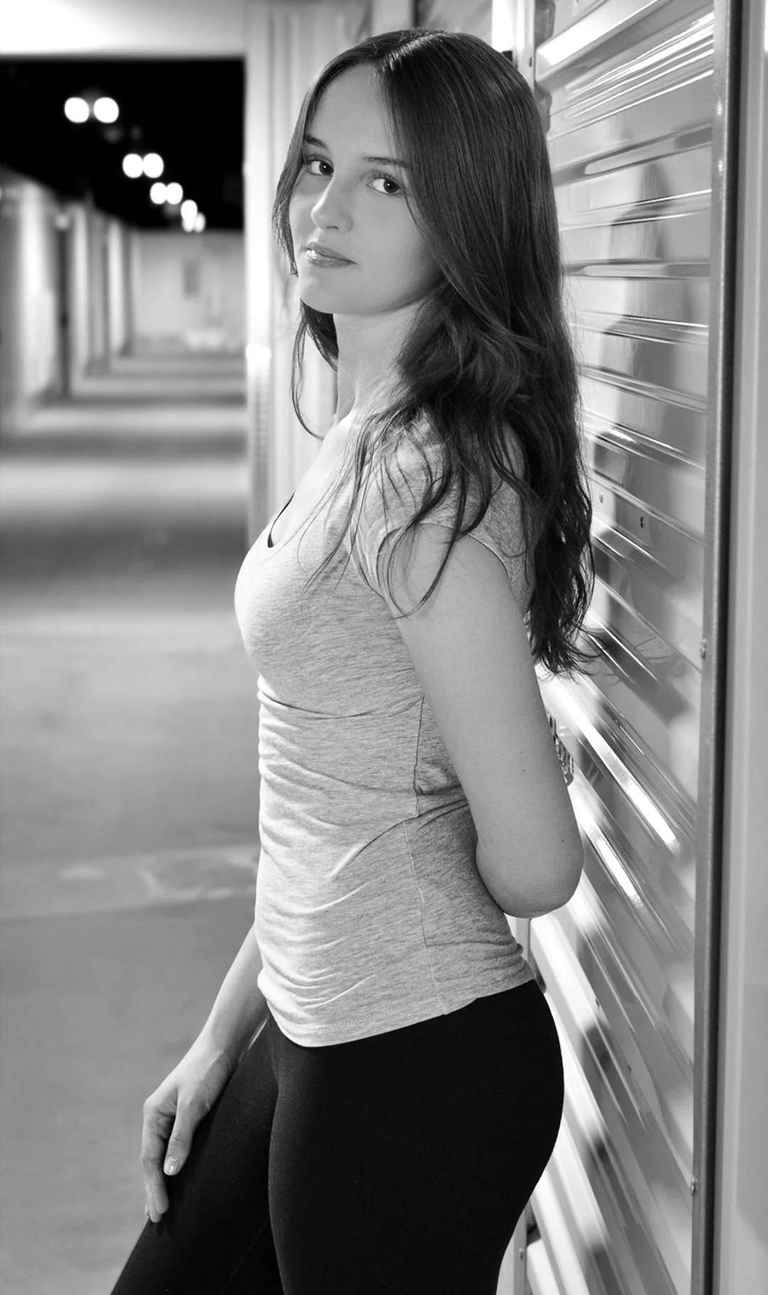 A woman with long dark hair leaning against a metal wall, wearing a fitted t-shirt and black pants, in a dimly lit indoor setting.