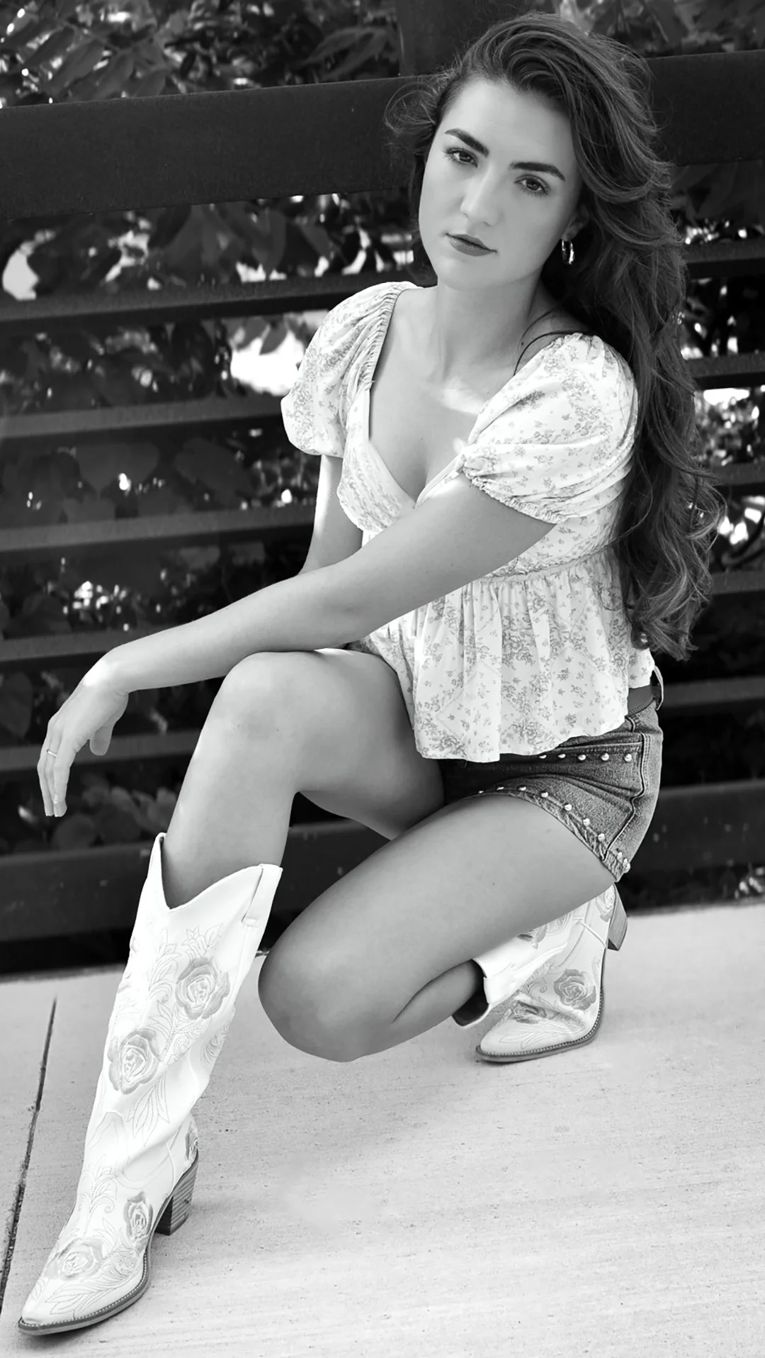A young woman with long wavy hair sitting outdoors on a bench, wearing a floral blouse, denim shorts, and cowboy boots.