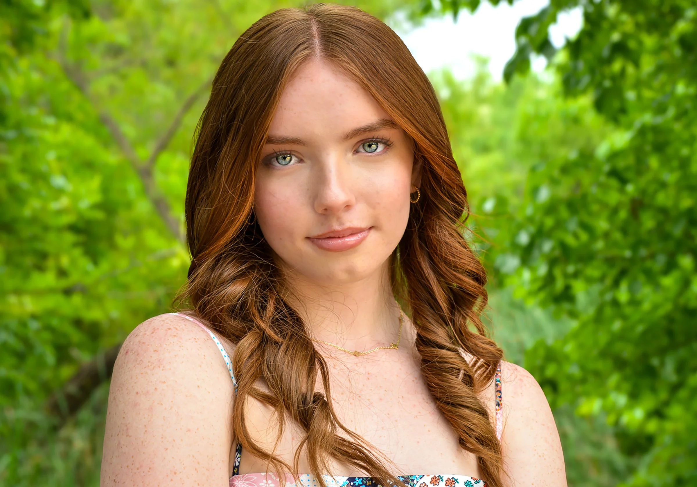 Close-up of a young woman with red hair and blue eyes, smiling softly, outdoors with green foliage in the background.