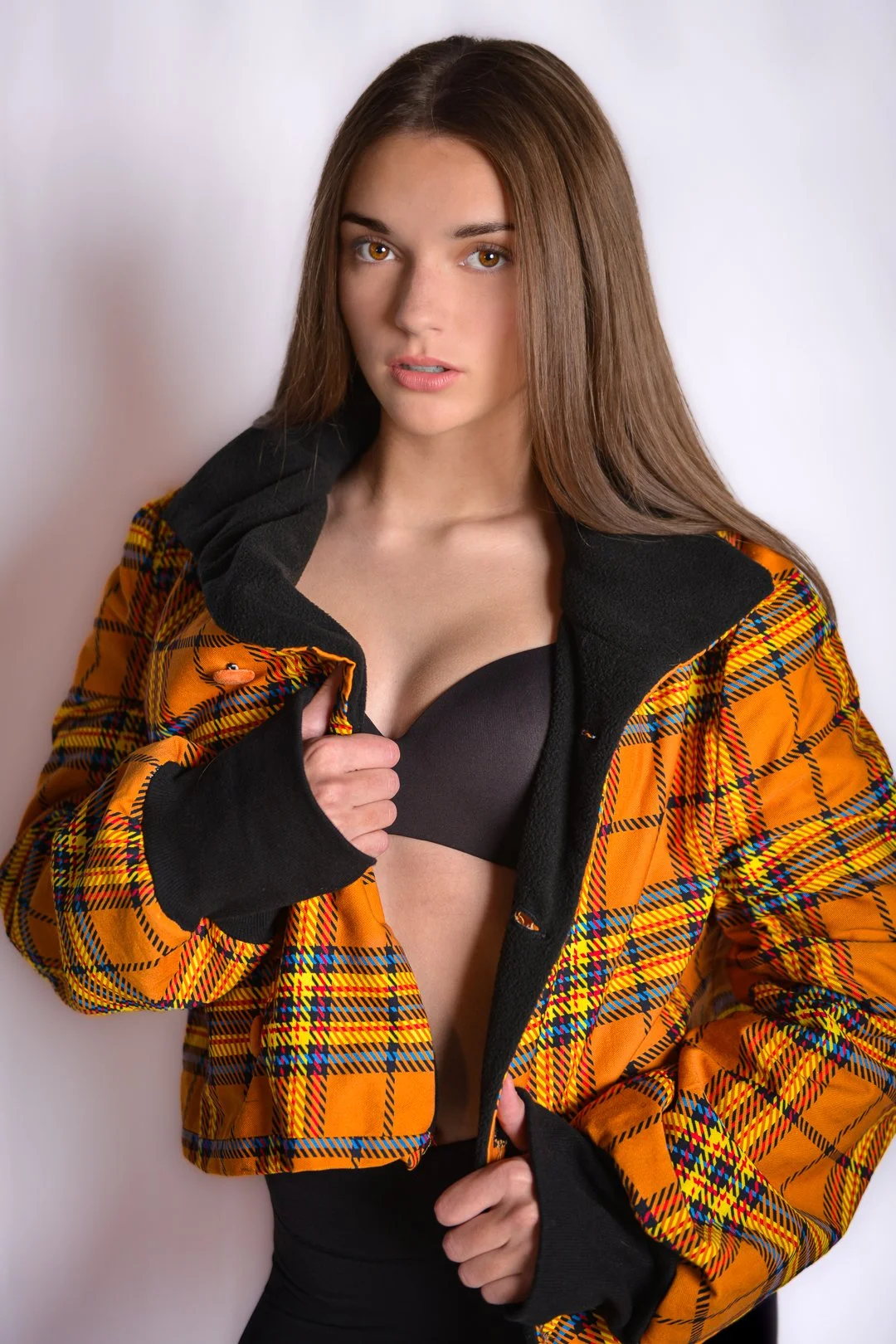 Young woman with long brown hair wearing a yellow plaid jacket and black bra, standing against a white background.