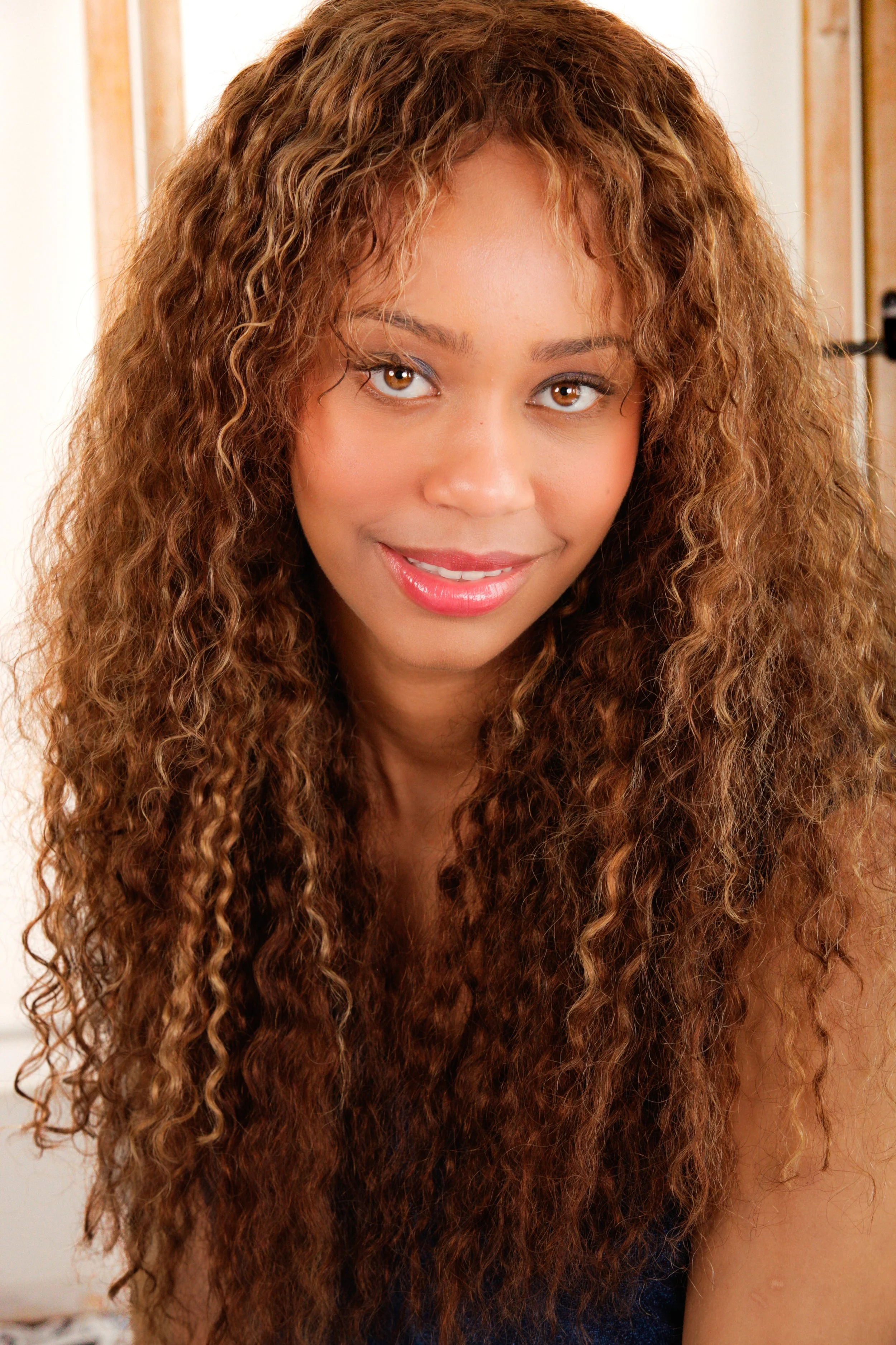 Close-up of a woman with long, curly auburn hair and brown eyes, smiling slightly, indoors with a wooden door and beige wall in background.