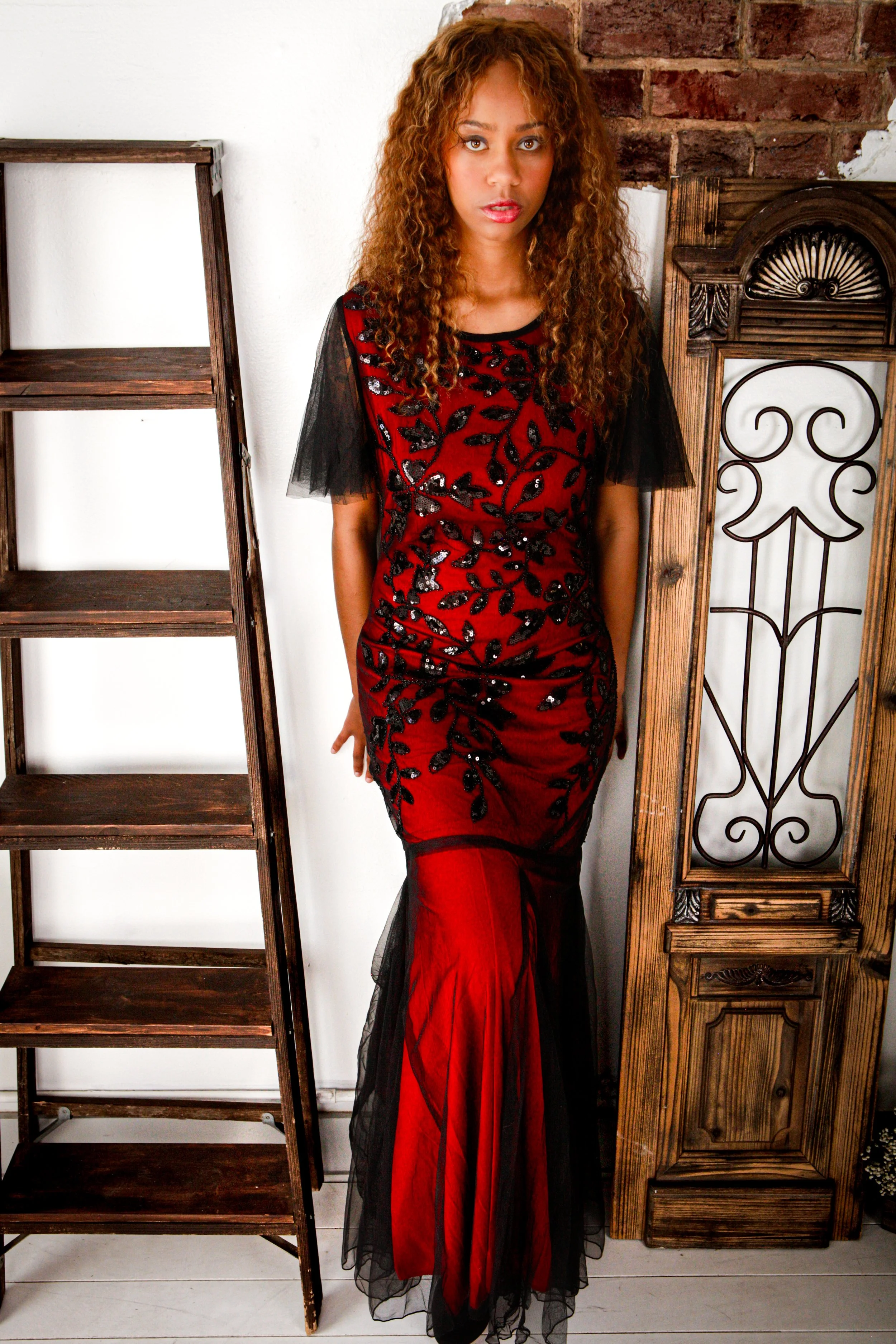 Woman with curly hair wearing a black and red dress with a sheer black overlay standing between a wooden ladder and a wooden screen, in front of a brick and white wall.