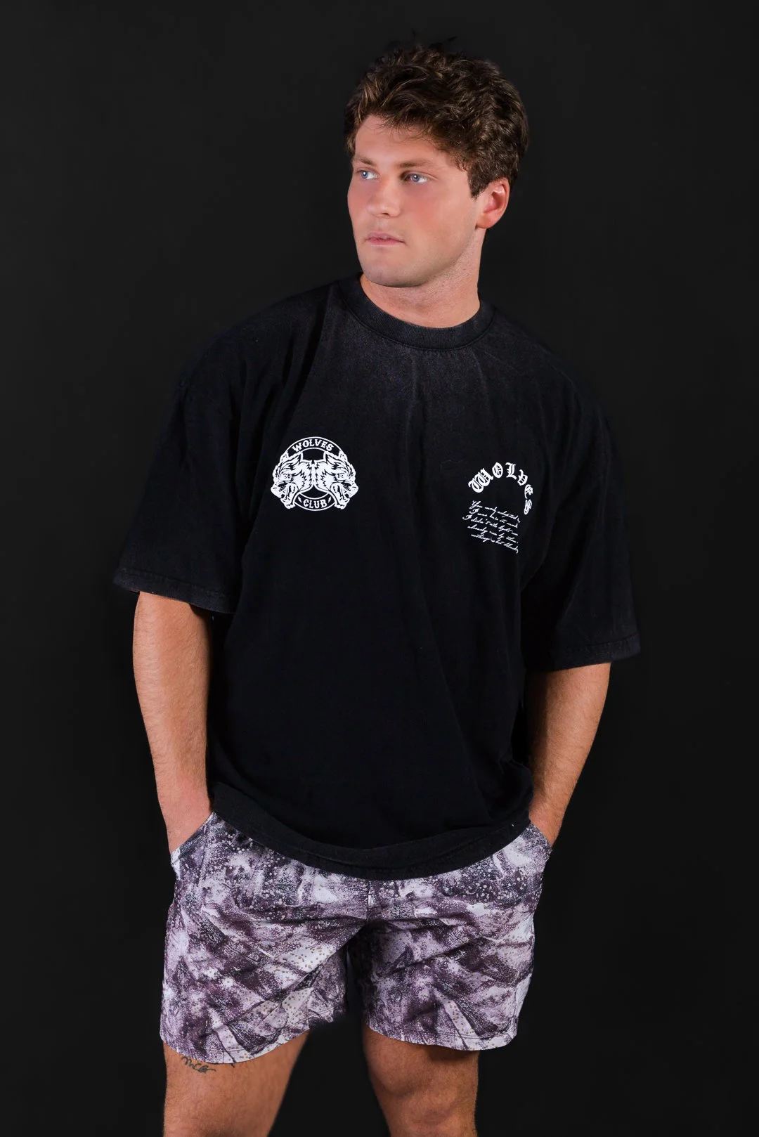 A young man with curly brown hair wearing a black t-shirt with white graphic logos and patterned shorts, standing against a black background.