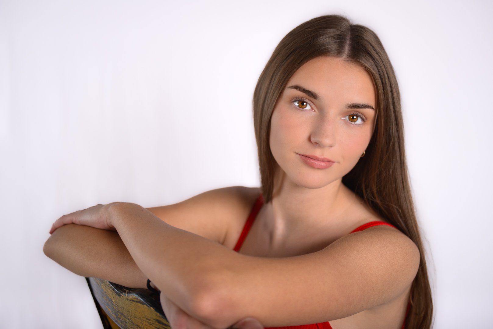 A young woman with long brown hair and brown eyes, wearing a red top, is smiling softly at the camera. She is posed with one arm resting on her shoulder against a plain white background.