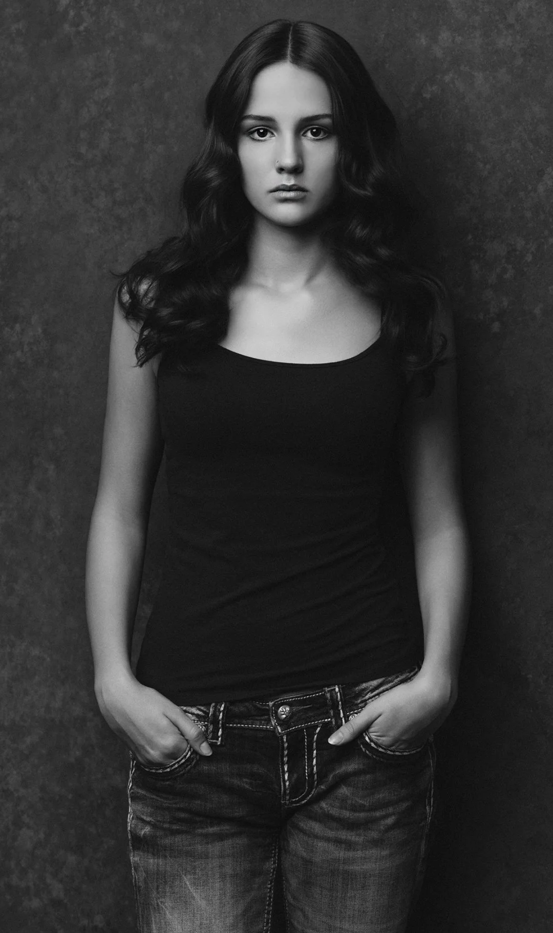 Black and white photo of a young woman with long dark hair, wearing a sleeveless top and jeans, standing against a textured dark background.