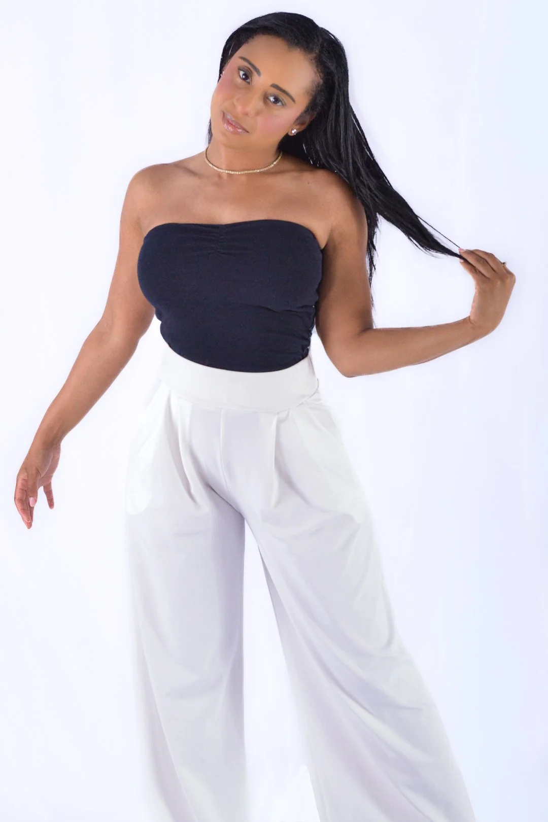 A woman wearing a black strapless top, white high-waisted pants, a pearl necklace, and earrings, posed against a white background, with one hand touching her hair.