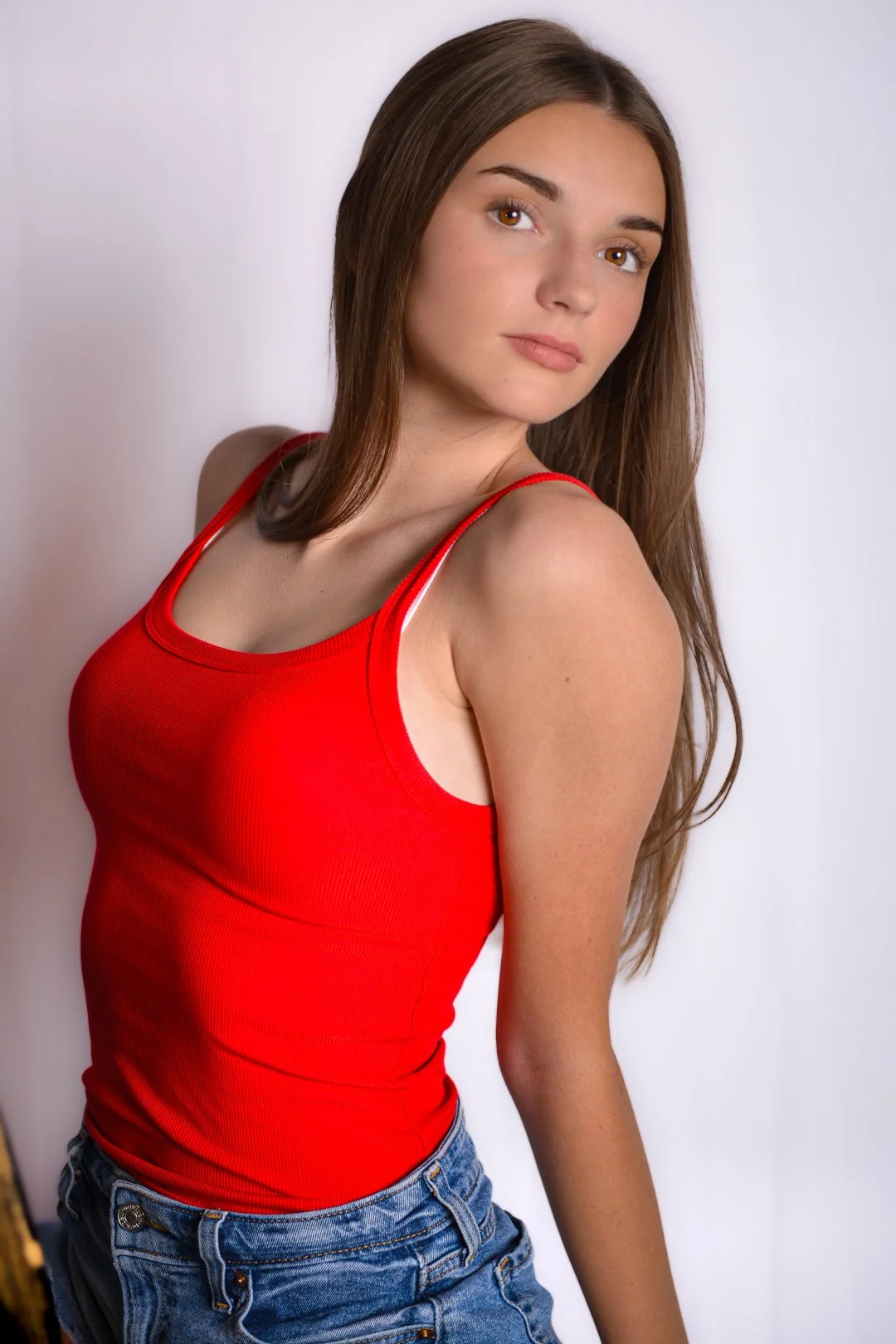A young woman with long brown hair and light skin wearing a red tank top and denim shorts, posing against a plain white background.