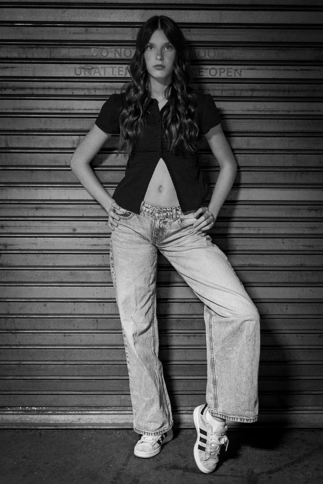 A young woman with long wavy hair standing in front of a closed metal door, wearing a black short-sleeve shirt, loose jeans, and sneakers, with her hands on her hips.