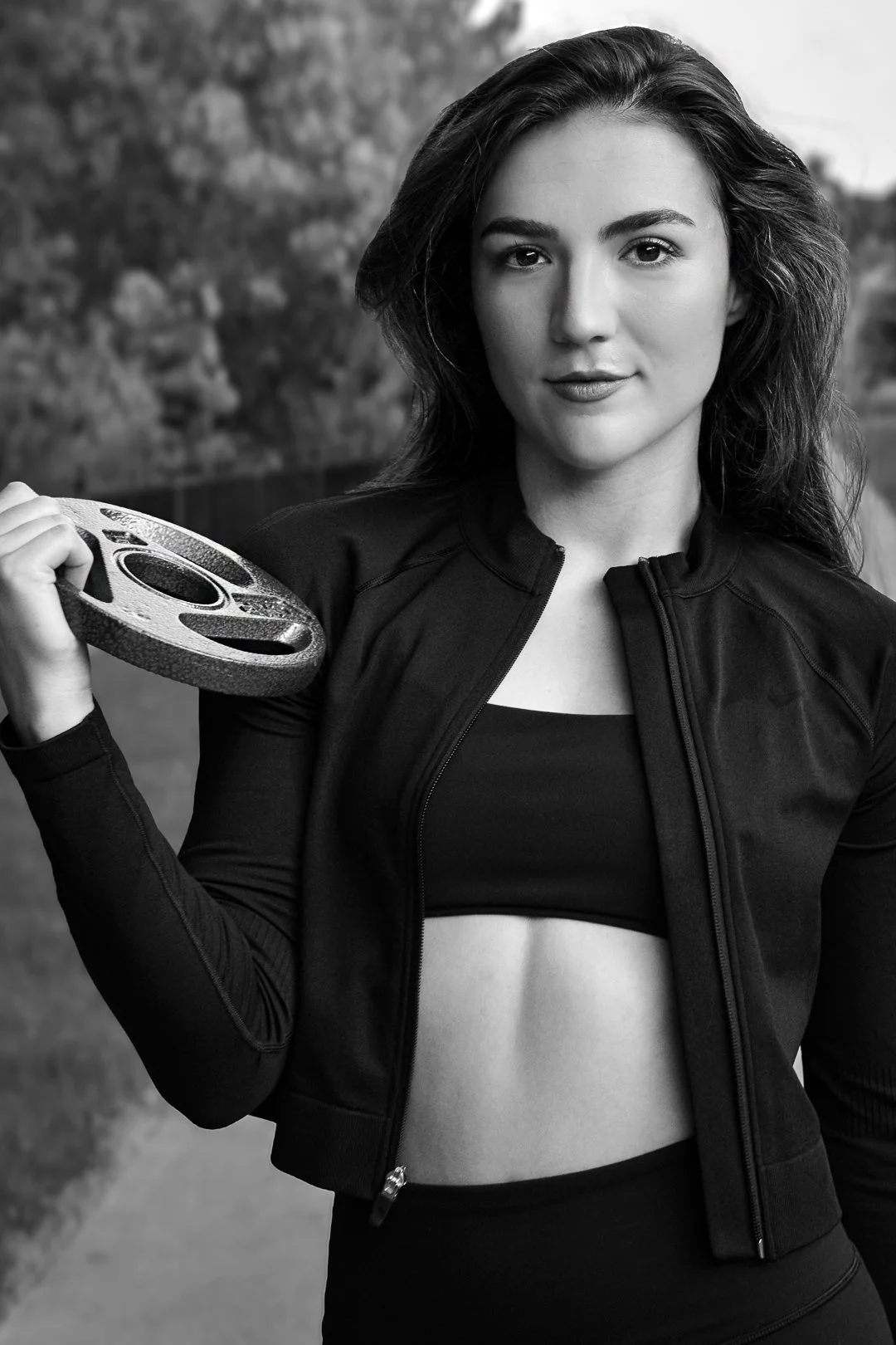 A young woman with dark hair and athletic clothing holds a weight plate over her shoulder outdoors.