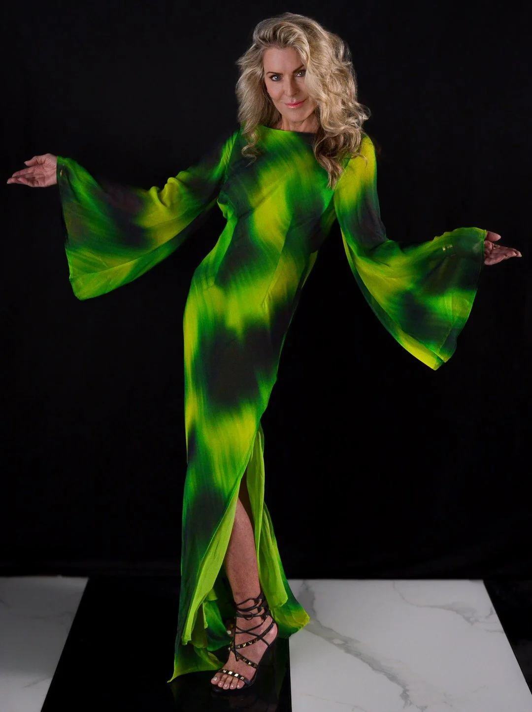 Woman in a flowing, long green dress with bell sleeves, standing on a black and white marble surface against a black background.