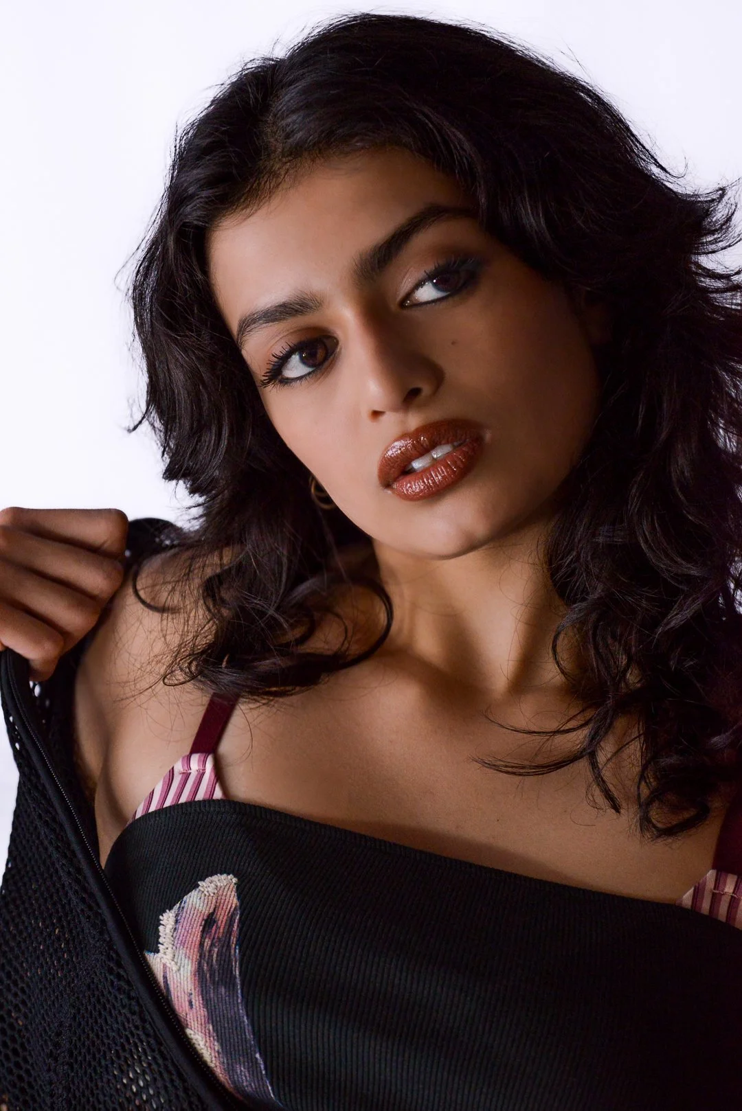 A woman with dark, wavy hair and makeup that emphasizes her eyes and lips, wearing a black top with a floral design, and holding a black jacket.